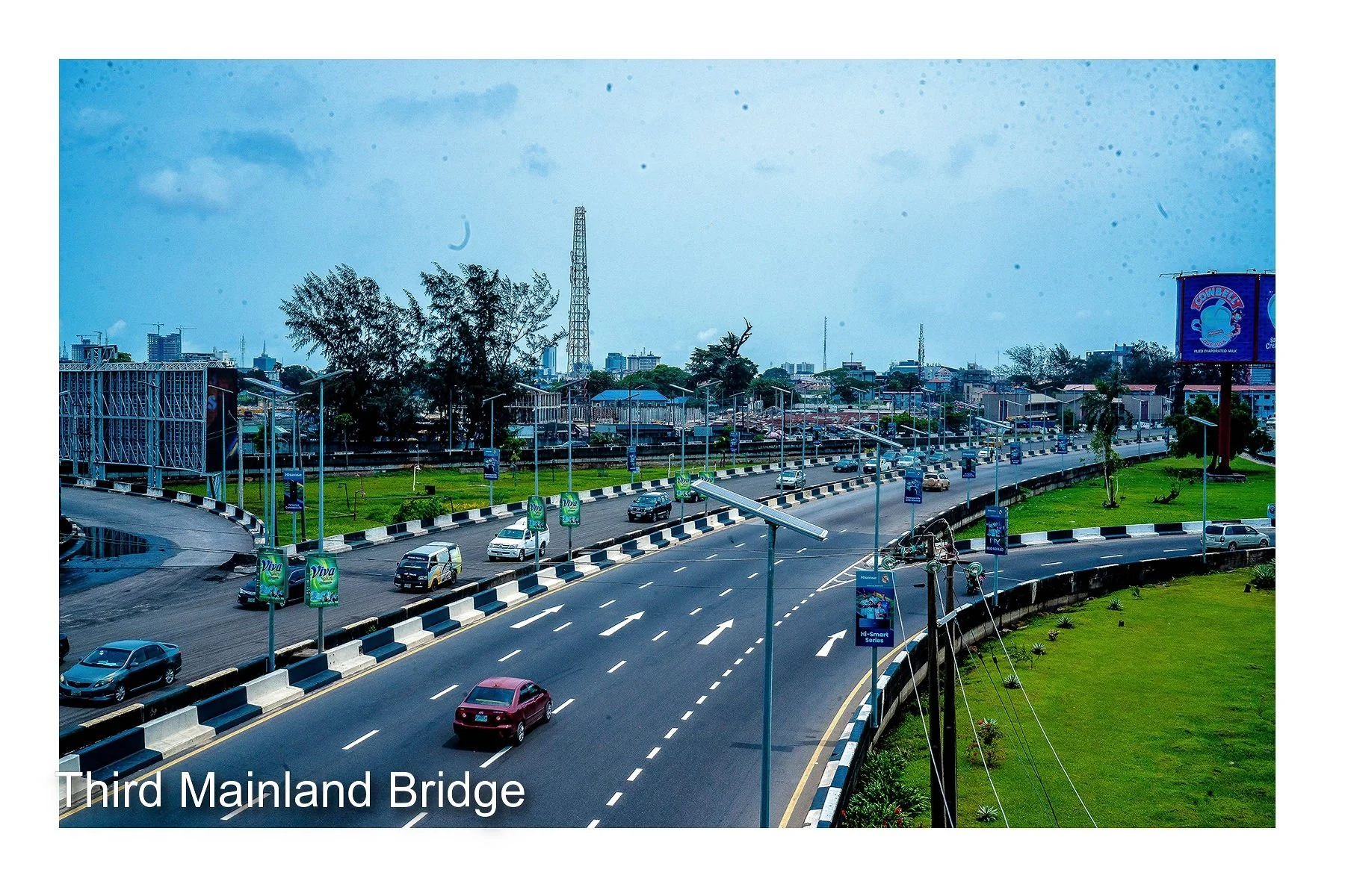 Photo of the Third Mainland Bridge in Nigeria showing multiple lanes of traffic, green grassy areas, street lamps, and buildings in the background under a partly cloudy sky.