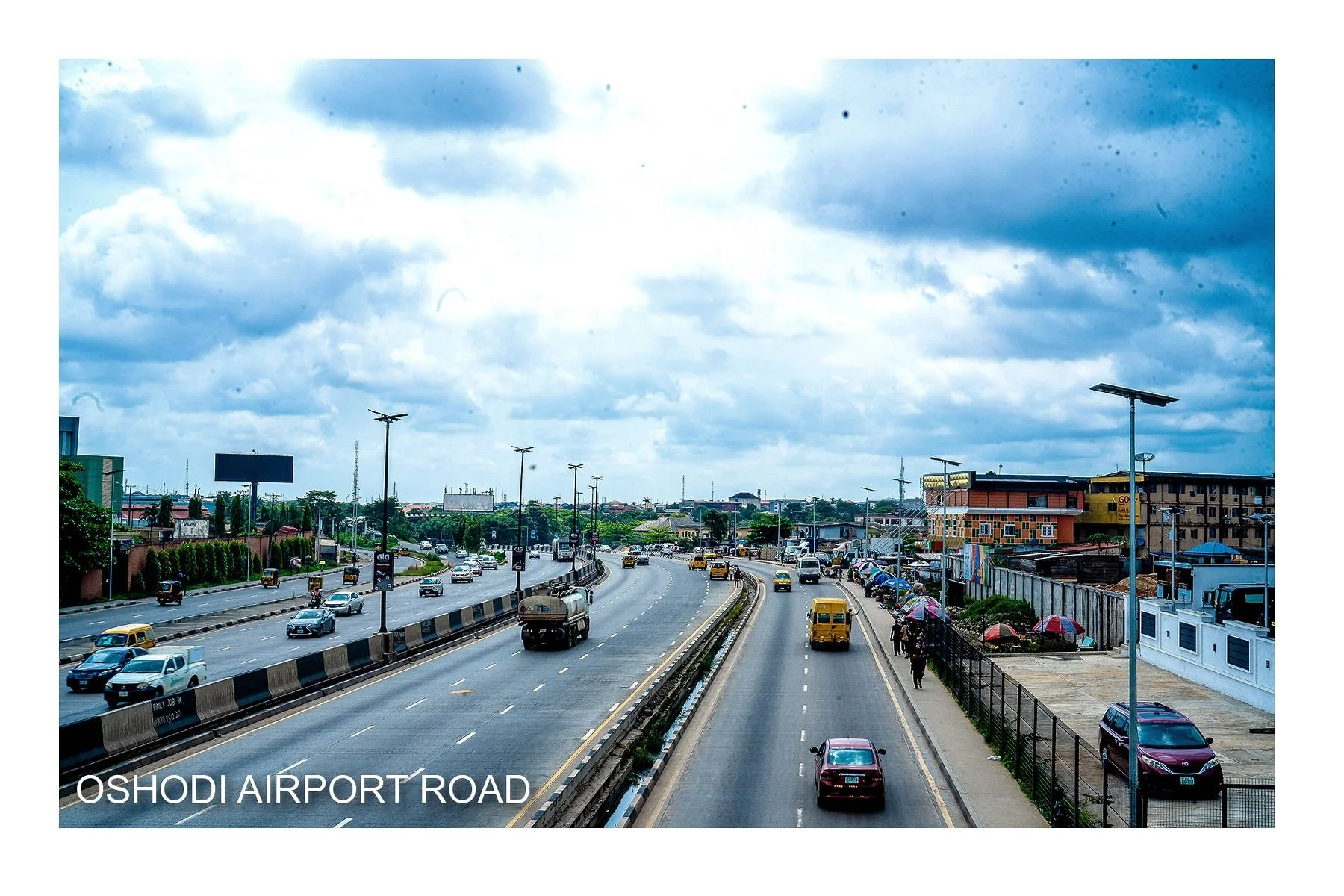 A busy urban street scene with cars, motorcycles, and buses, under a cloudy sky, seen from an elevated position on Oshodi Airport Road in Lagos, Nigeria.