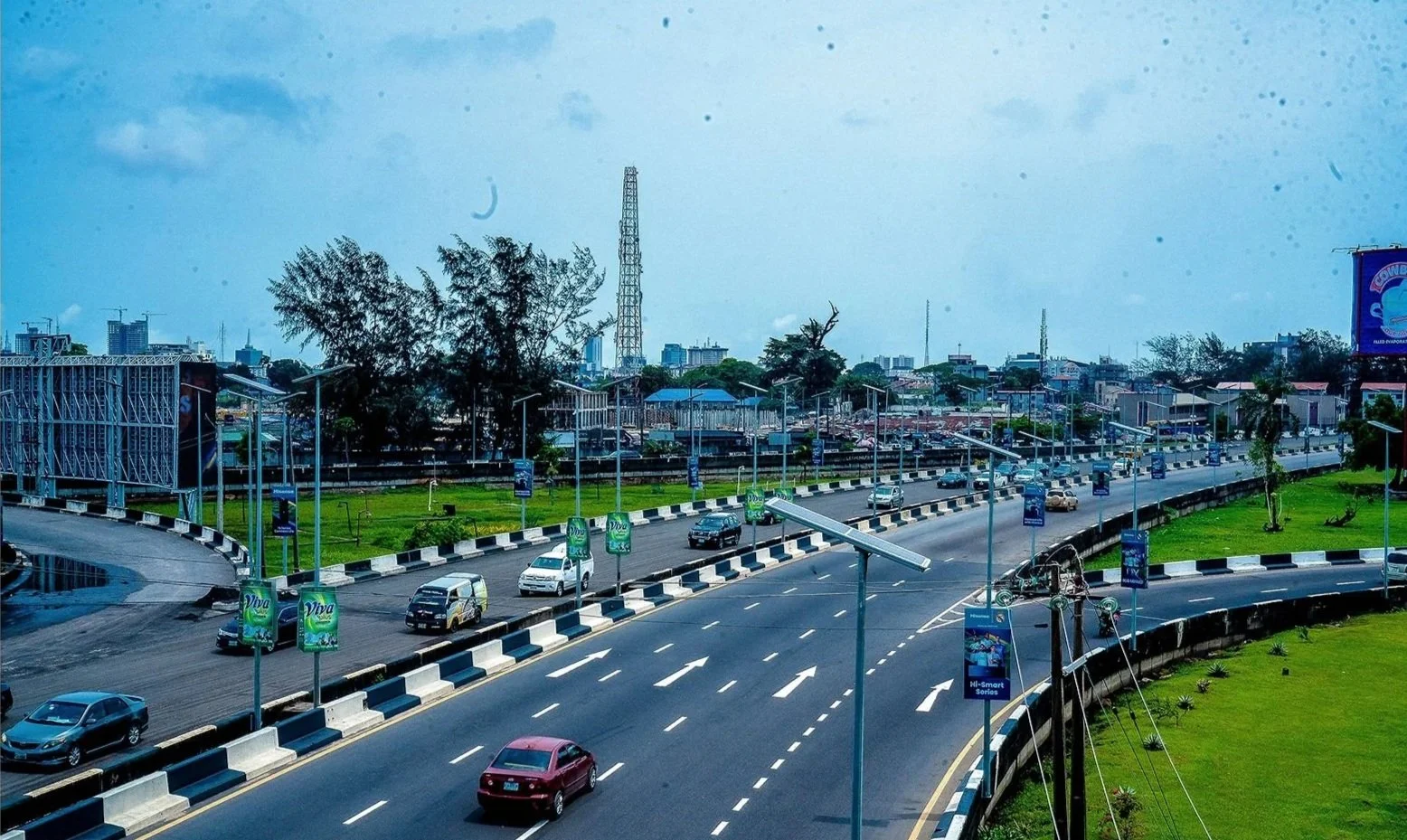 City view of a highway with cars, streetlights, billboards, green grass, trees, and a background of buildings and a communication tower under a partly cloudy sky.