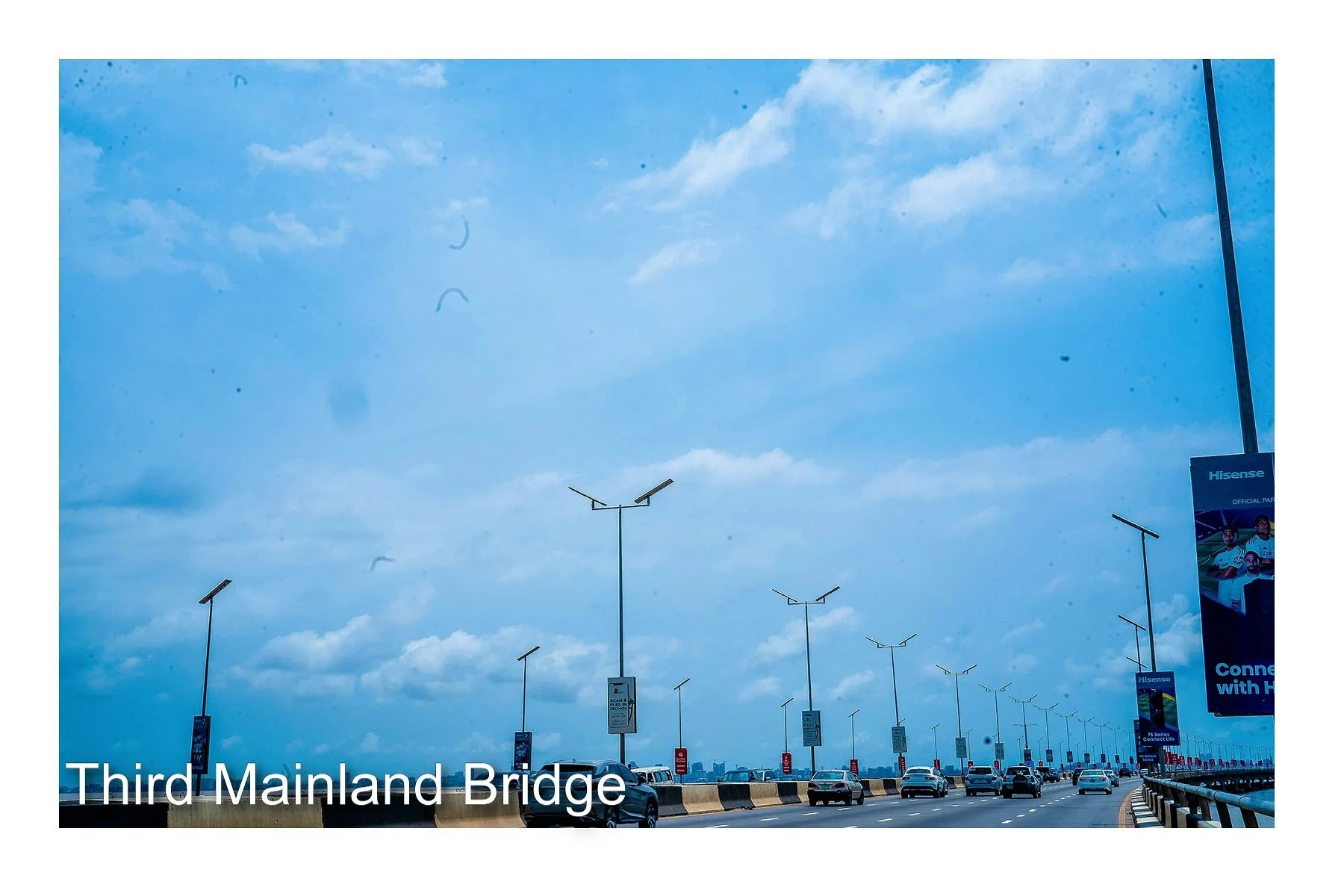 A view of the Third Mainland Bridge with multiple cars driving on it, lined with streetlights and advertisement banners, under a partly cloudy sky.