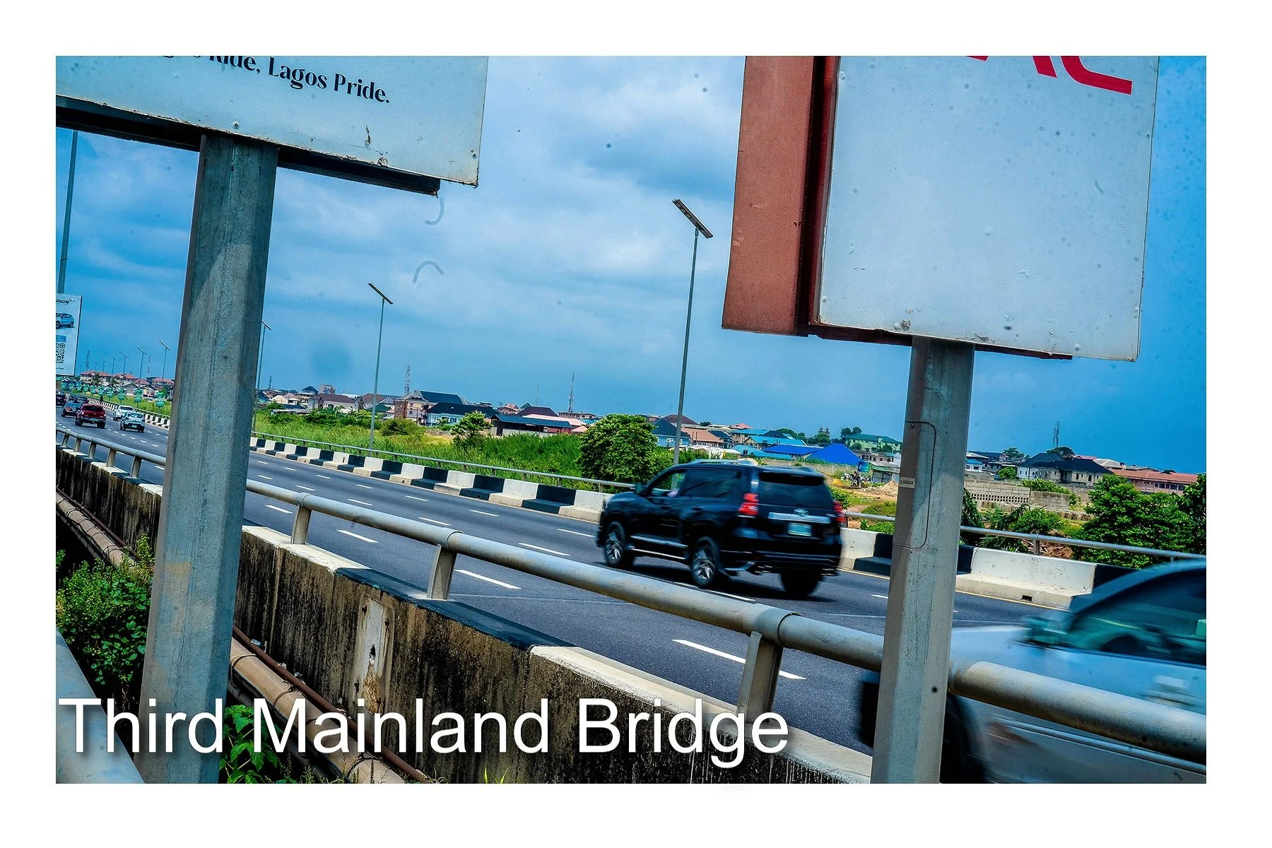 View of the Third Mainland Bridge with cars driving over it, and some billboards visible in the foreground, with a cloudy sky and buildings in the background.