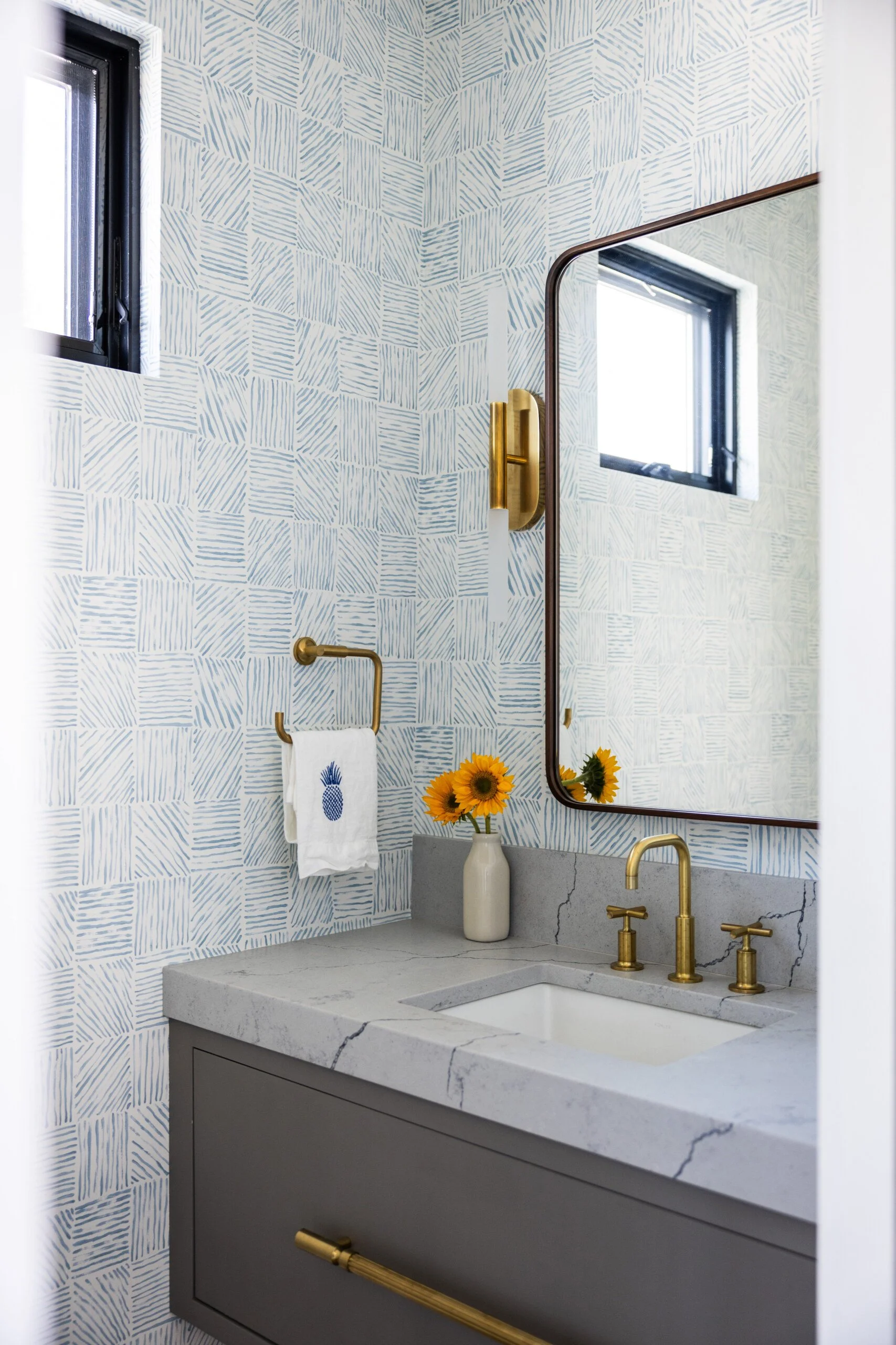 A modern bathroom with a geometric patterned wallpaper, a large rectangular mirror, gold fixtures, a small window, a gray vanity with a white sink, and a vase of sunflowers on the countertop.