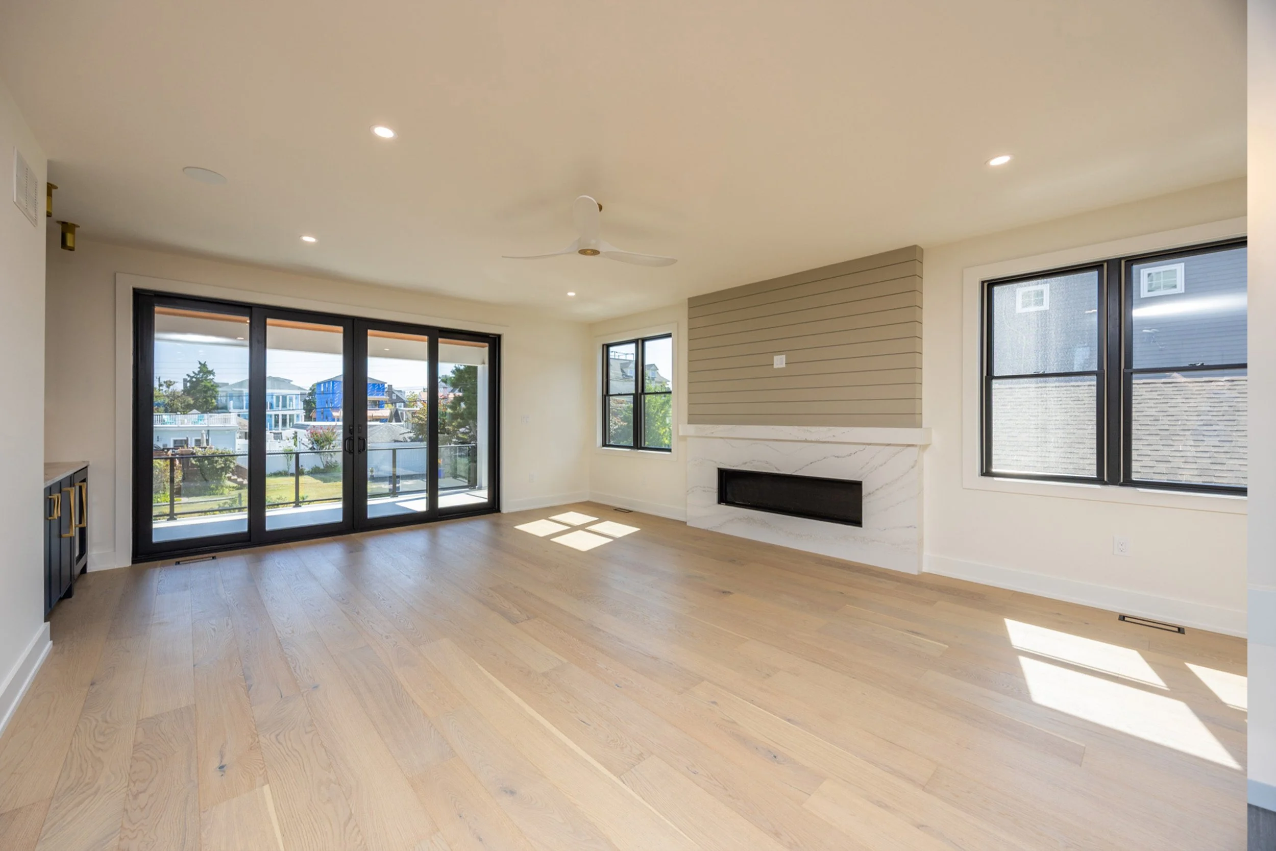 Empty living room with large sliding glass doors leading to a balcony, hardwood flooring, windows letting in natural light, and a modern fireplace with a beige and white marble finish.