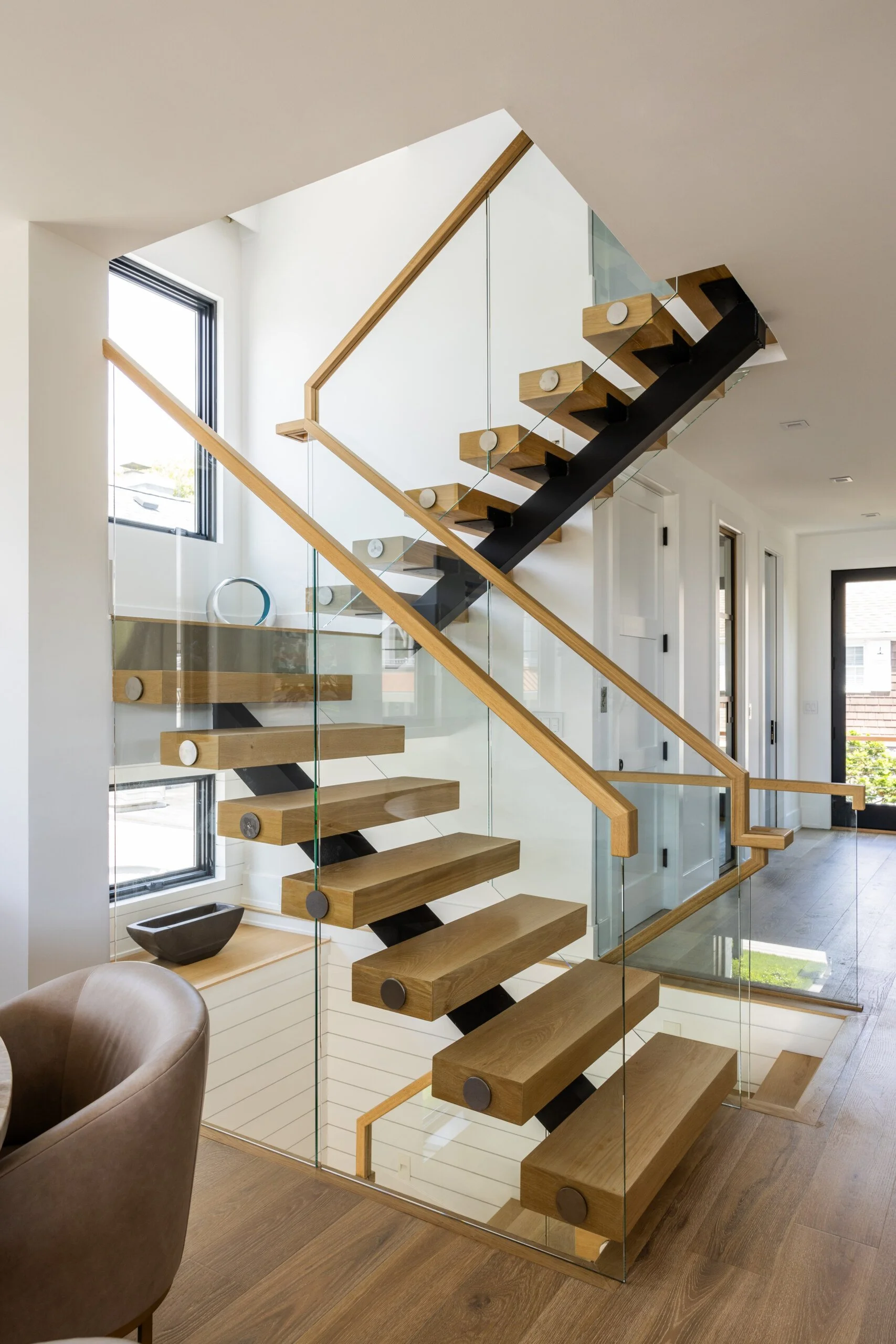 Modern wooden staircase with glass sides in a bright indoor space with large windows.