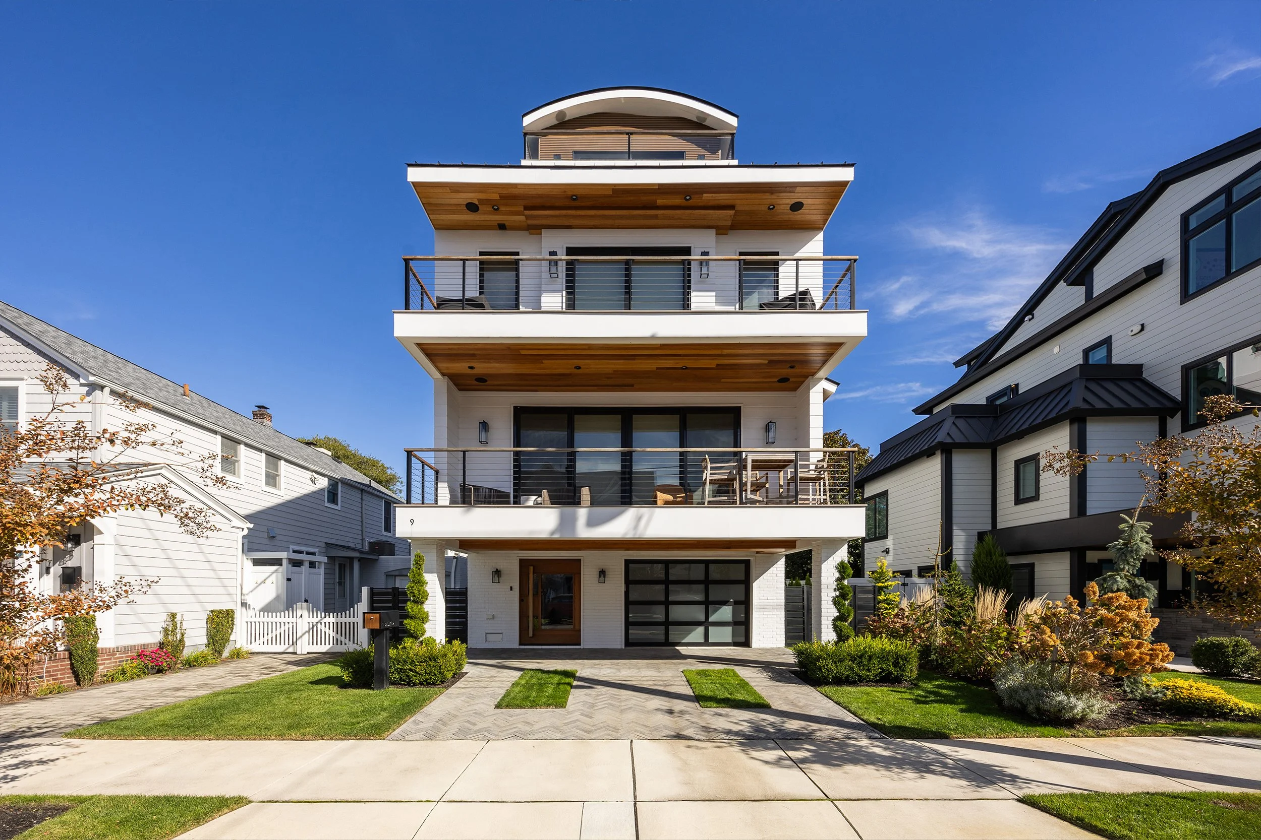 Modern multi-story house with white exterior, large glass doors, and balconies with black railings, surrounded by well-maintained lawn and landscaping, under a clear blue sky.