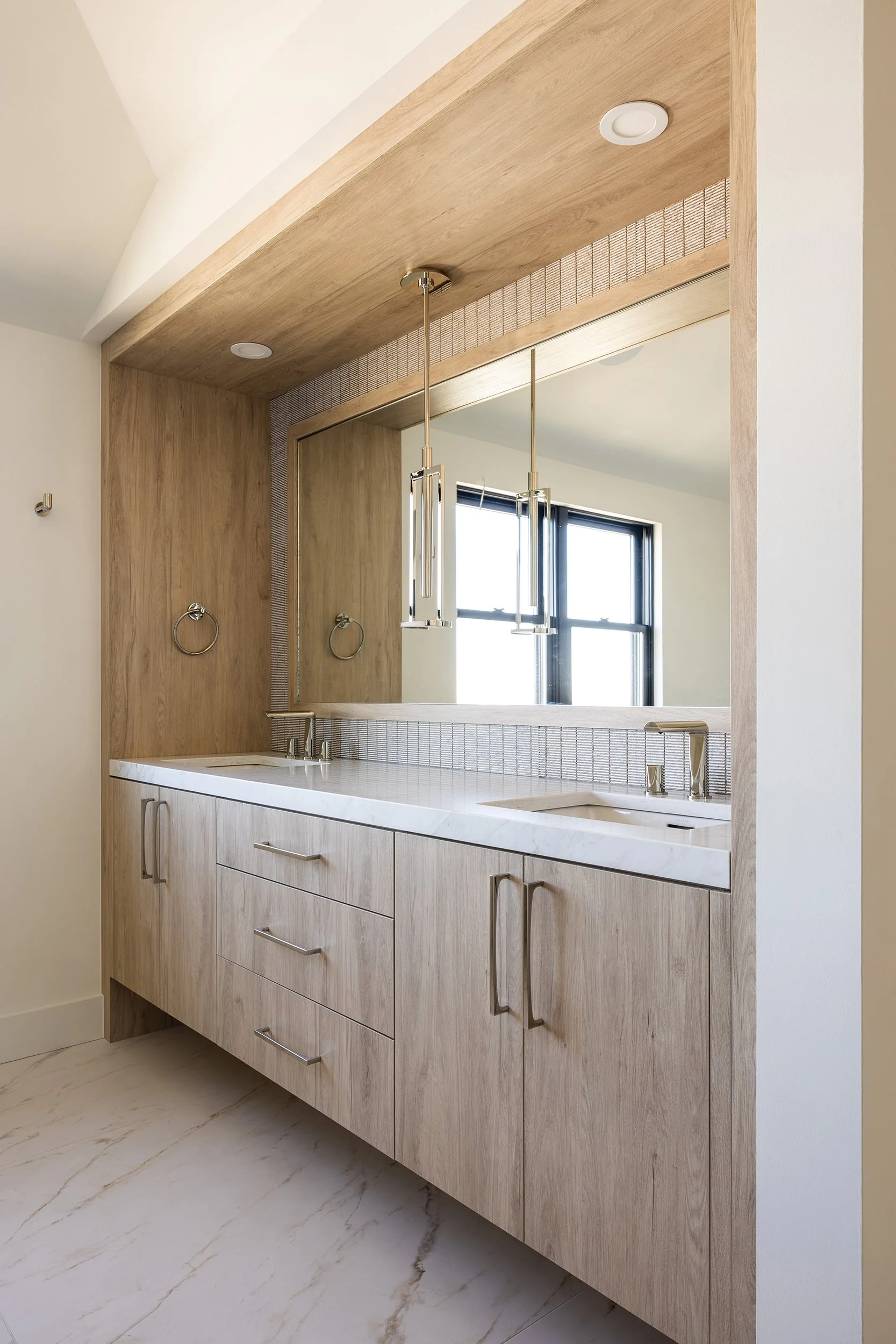 Modern bathroom vanity with light wood cabinets, white countertop, large mirror, two hanging pendant lights, with two windows reflected in the mirror.