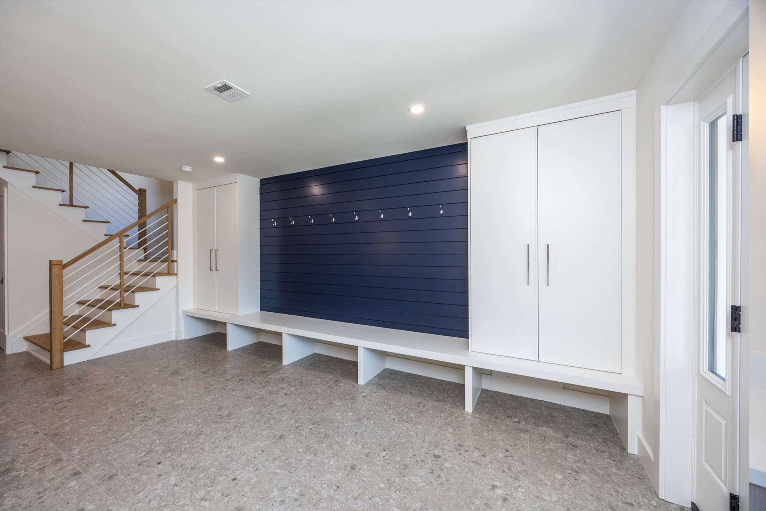 Empty entryway with built-in storage bench, white cabinetry, and a navy blue accent wall with hooks, near a staircase.