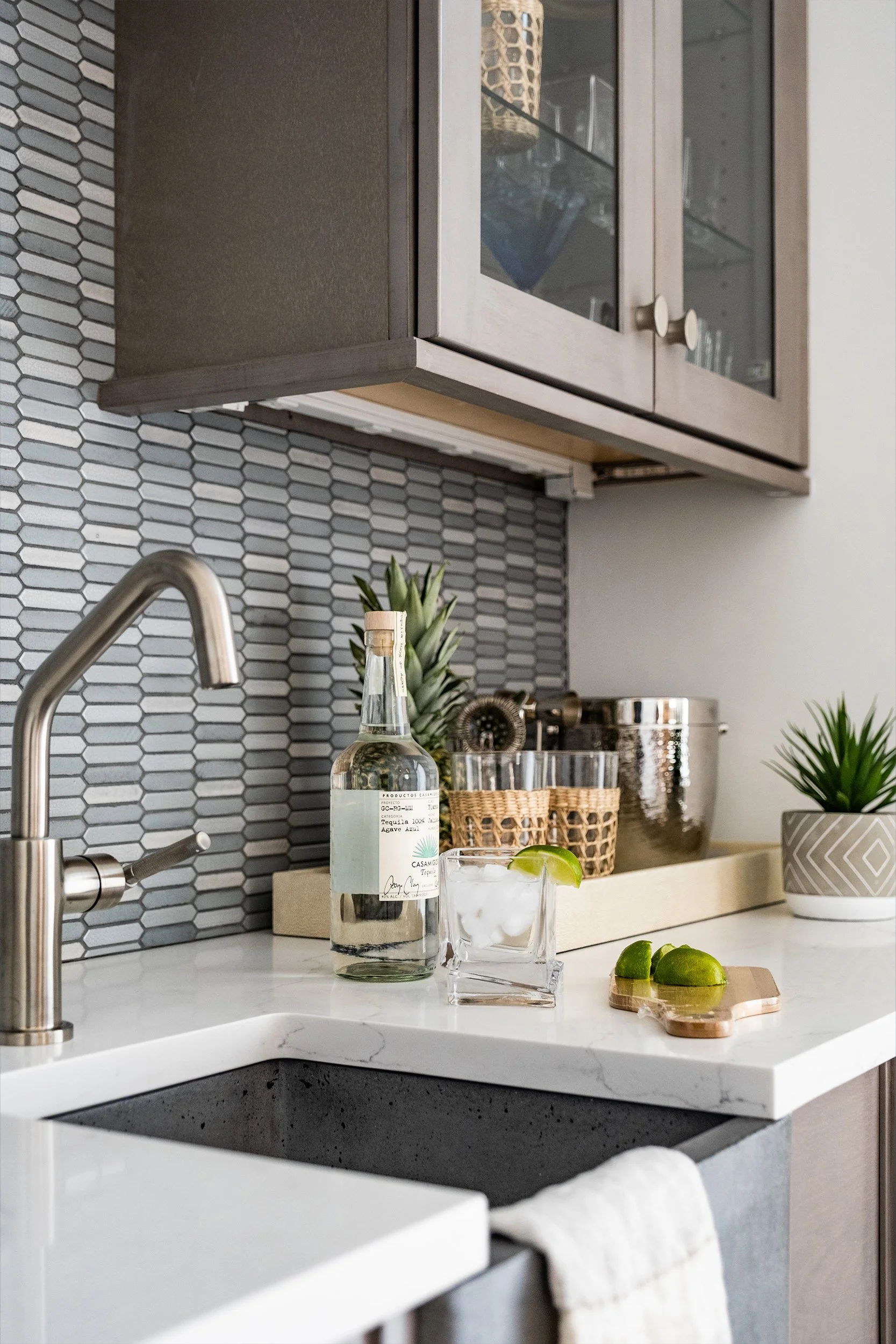 Modern kitchen counter with a bottle of tequila, a glass with ice and lime, fresh lime wedges on a small cutting board, pineapple and potted plant in the background.