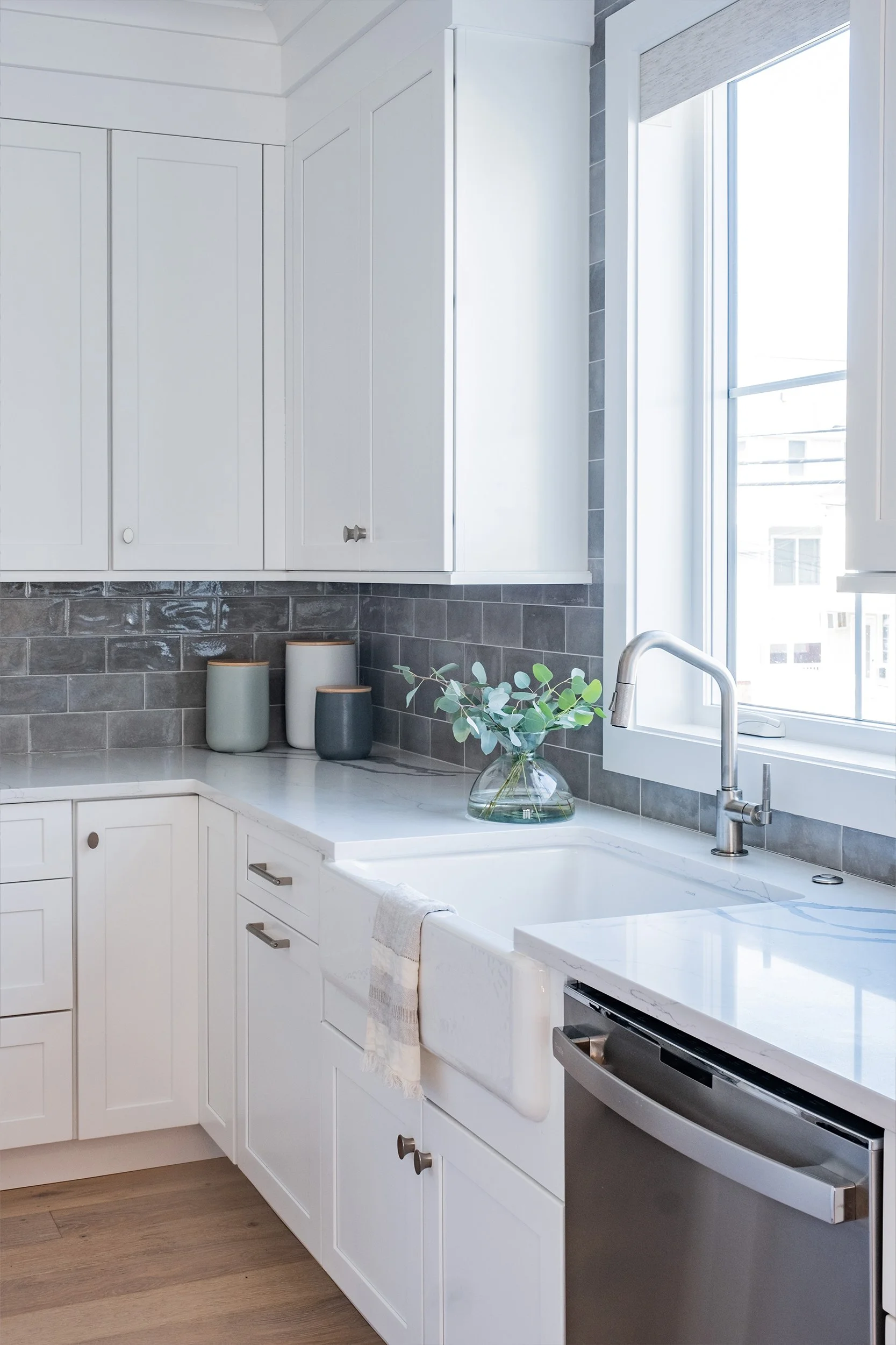 Modern kitchen with white cabinets, gray tile backsplash, marble countertop, a window above the sink, glass vase with greenery, and three ceramic containers on the counter.