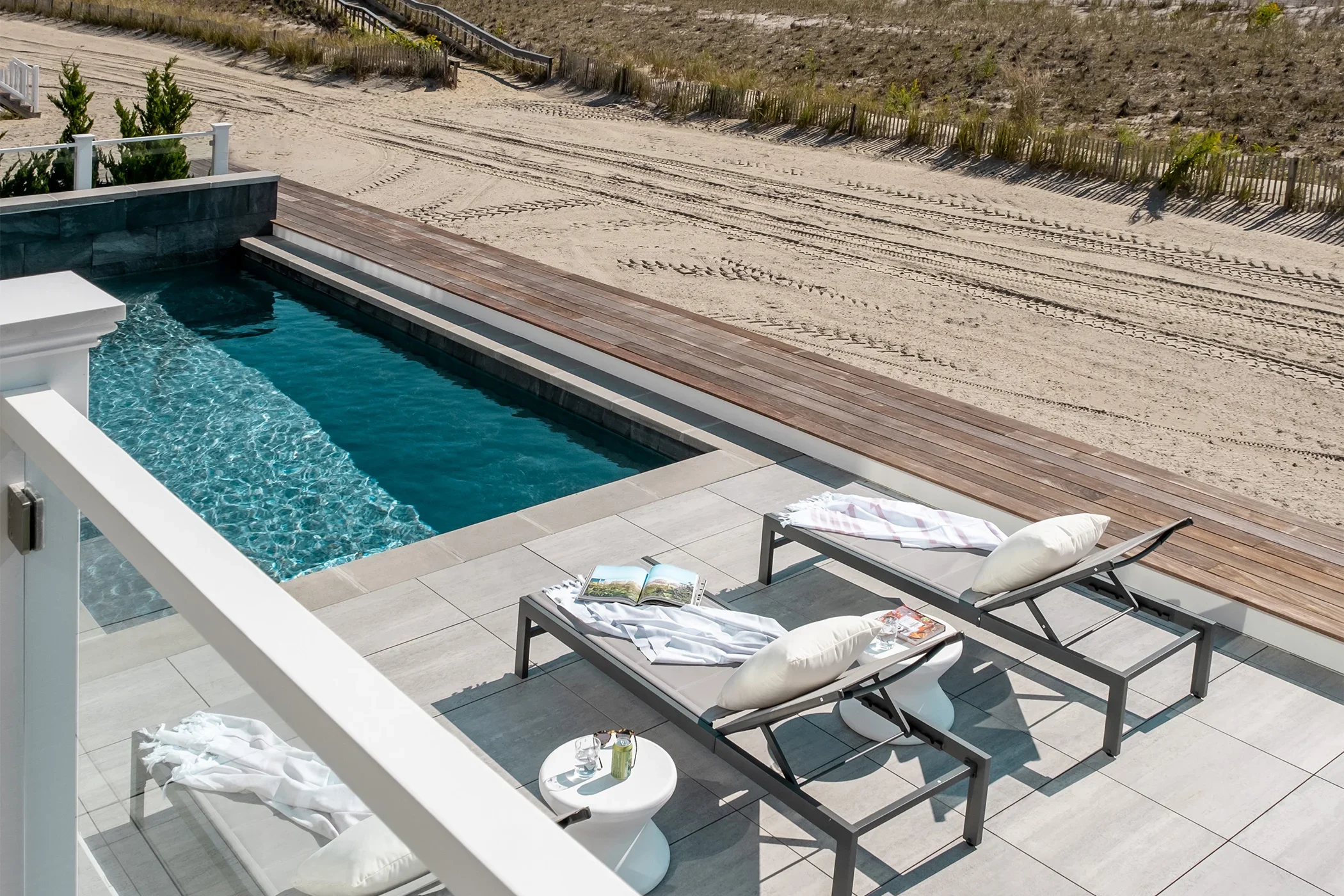 Empty poolside area with two lounge chairs, pillows, a magazine, and a small side table on a patio near a sandy beach.