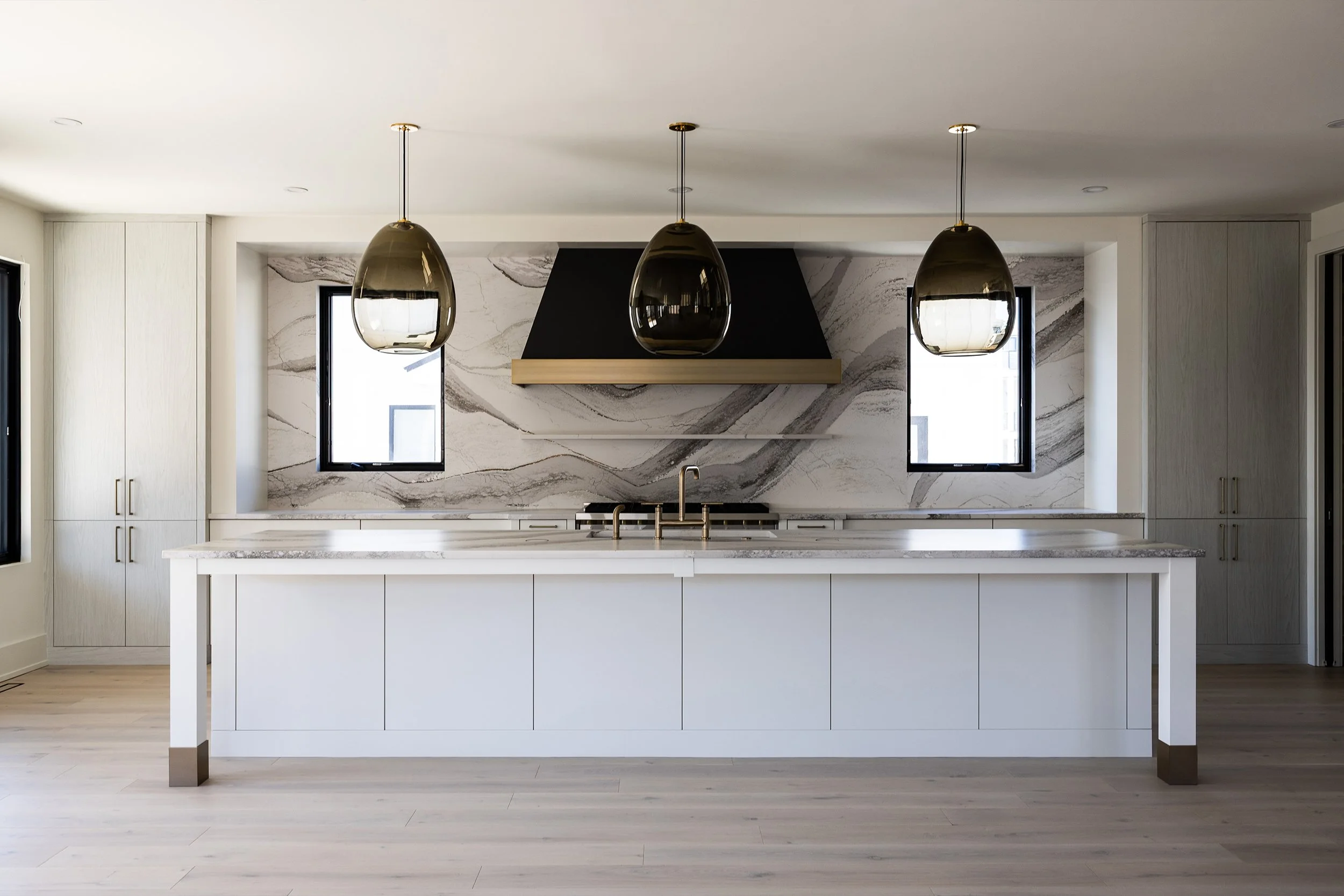 Modern kitchen with white island, black range hood, marble backsplash, pendant lights, and two windows.