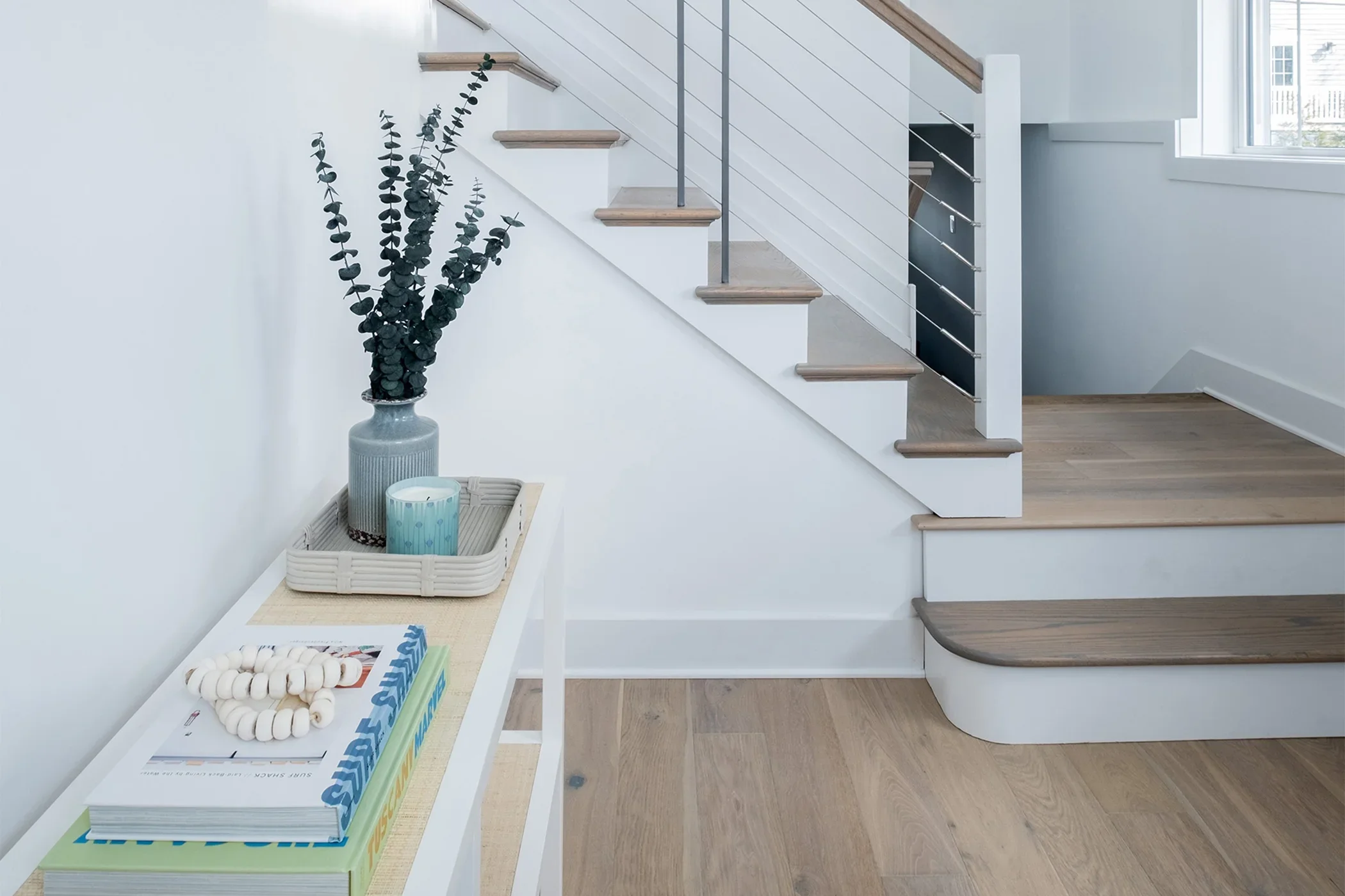 Indoor scene with a white table holding a coiled white beaded decoration, a stack of magazines, and a tray with a striped candle and a blue candle. A tall gray vase with eucalyptus branches is on top of the table. In the background, wooden stairs with metal railing lead upstairs, with a window on the right side.