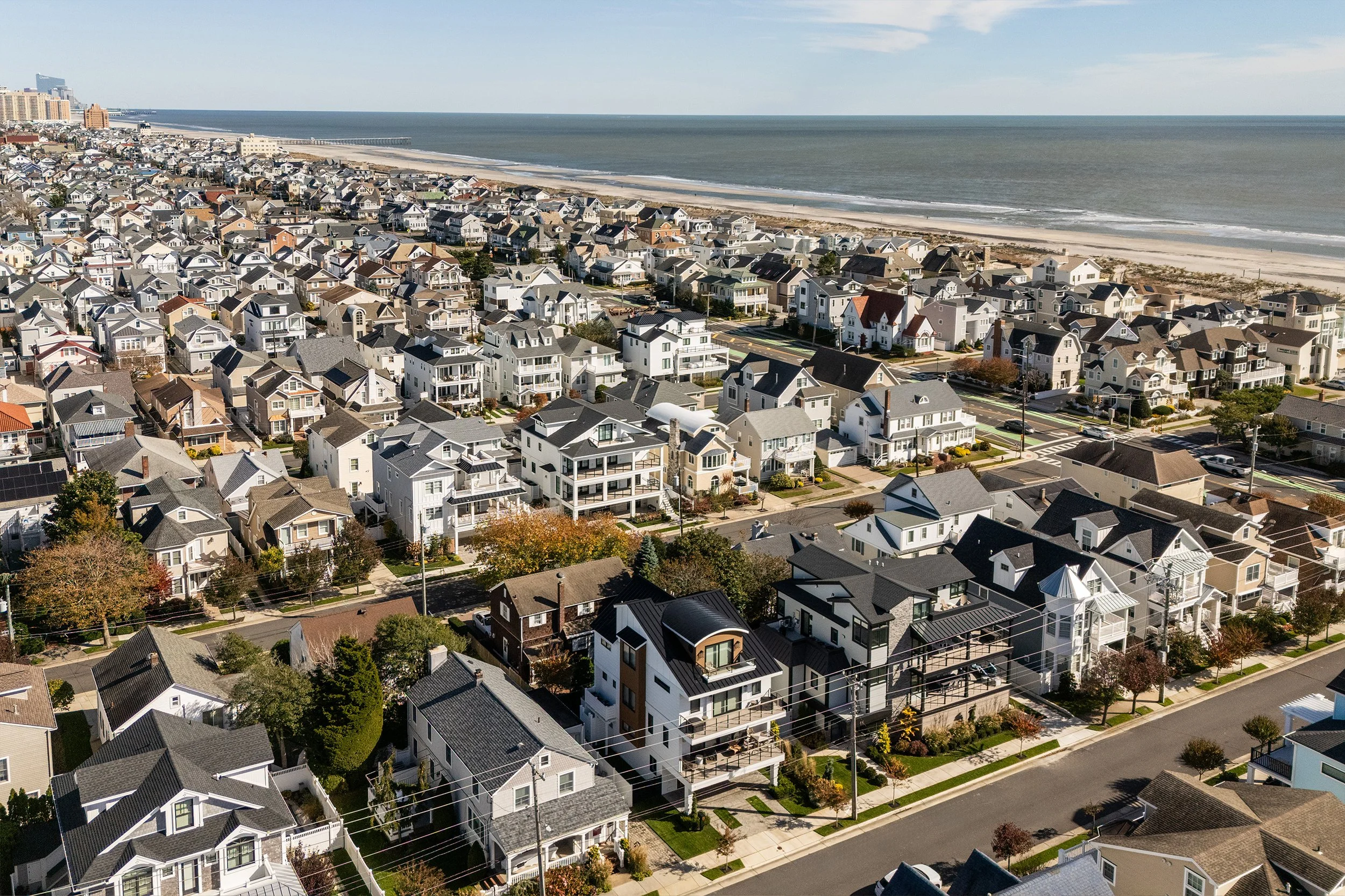 Aerial view of a coastal neighborhood with numerous houses facing the ocean, showing a mix of modern and traditional architecture, with some houses featuring balconies, greenery, and trees lining the streets.