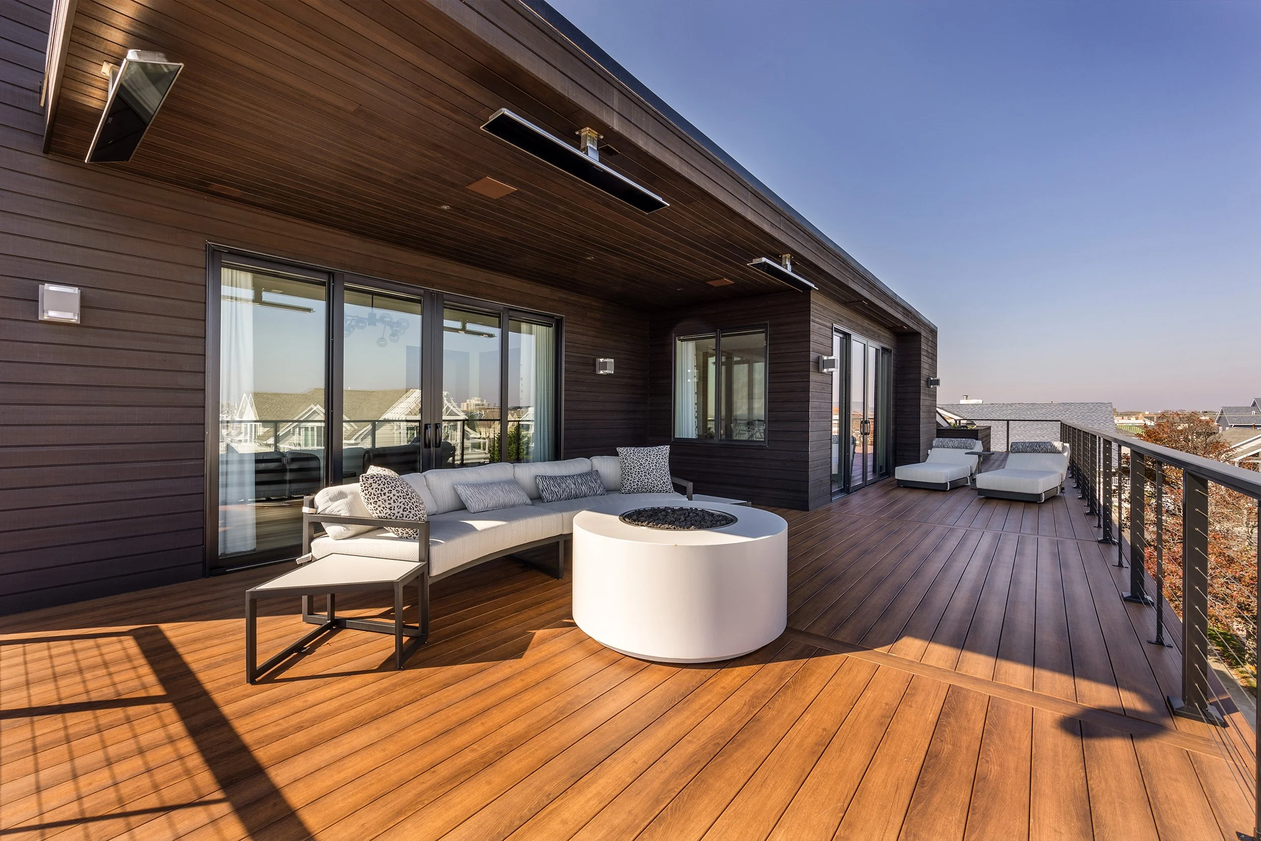 Spacious rooftop balcony with wooden flooring, a white outdoor sofa with cushions, lounge chairs, and a modern fire pit, surrounded by a black railing, under a clear blue sky.