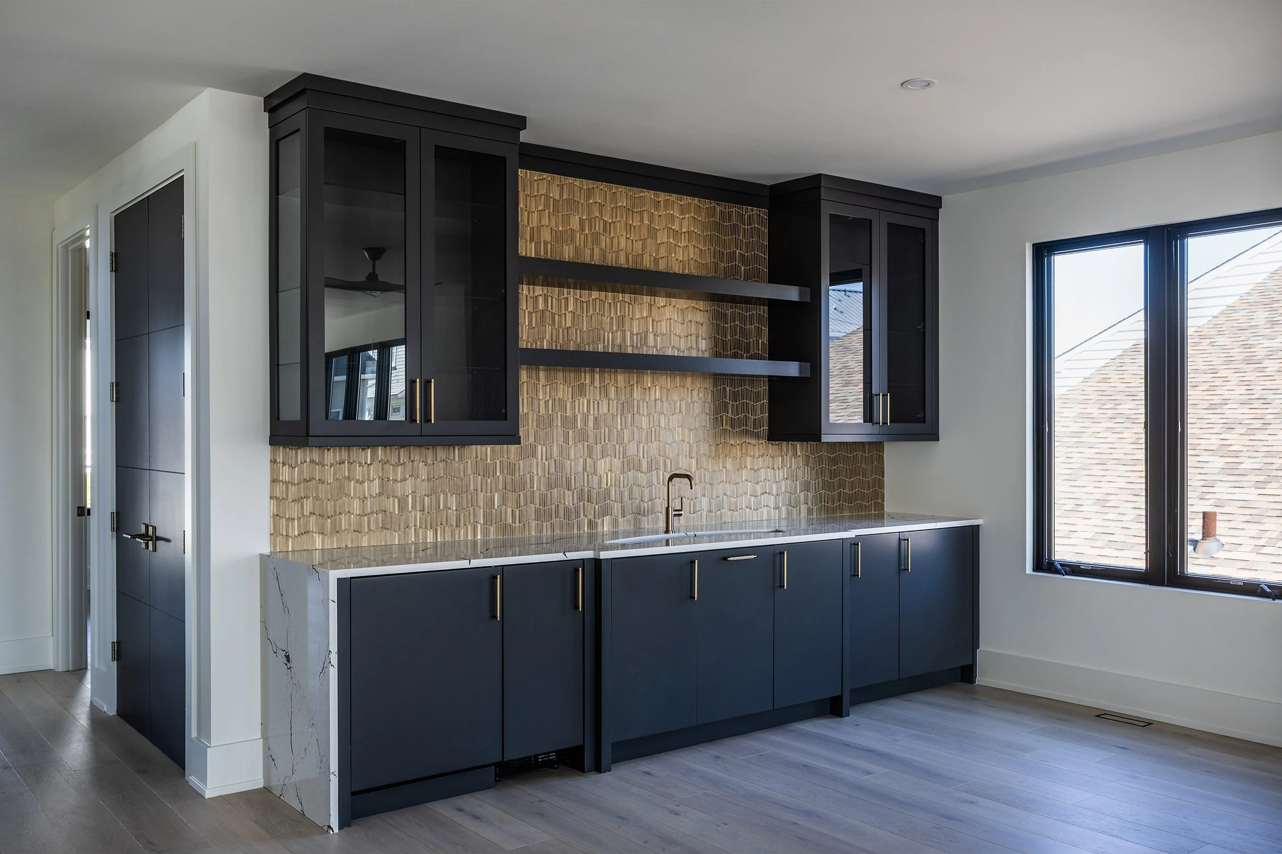 Modern kitchen with black cabinets, gold textured backsplash, and open shelving near a large window.
