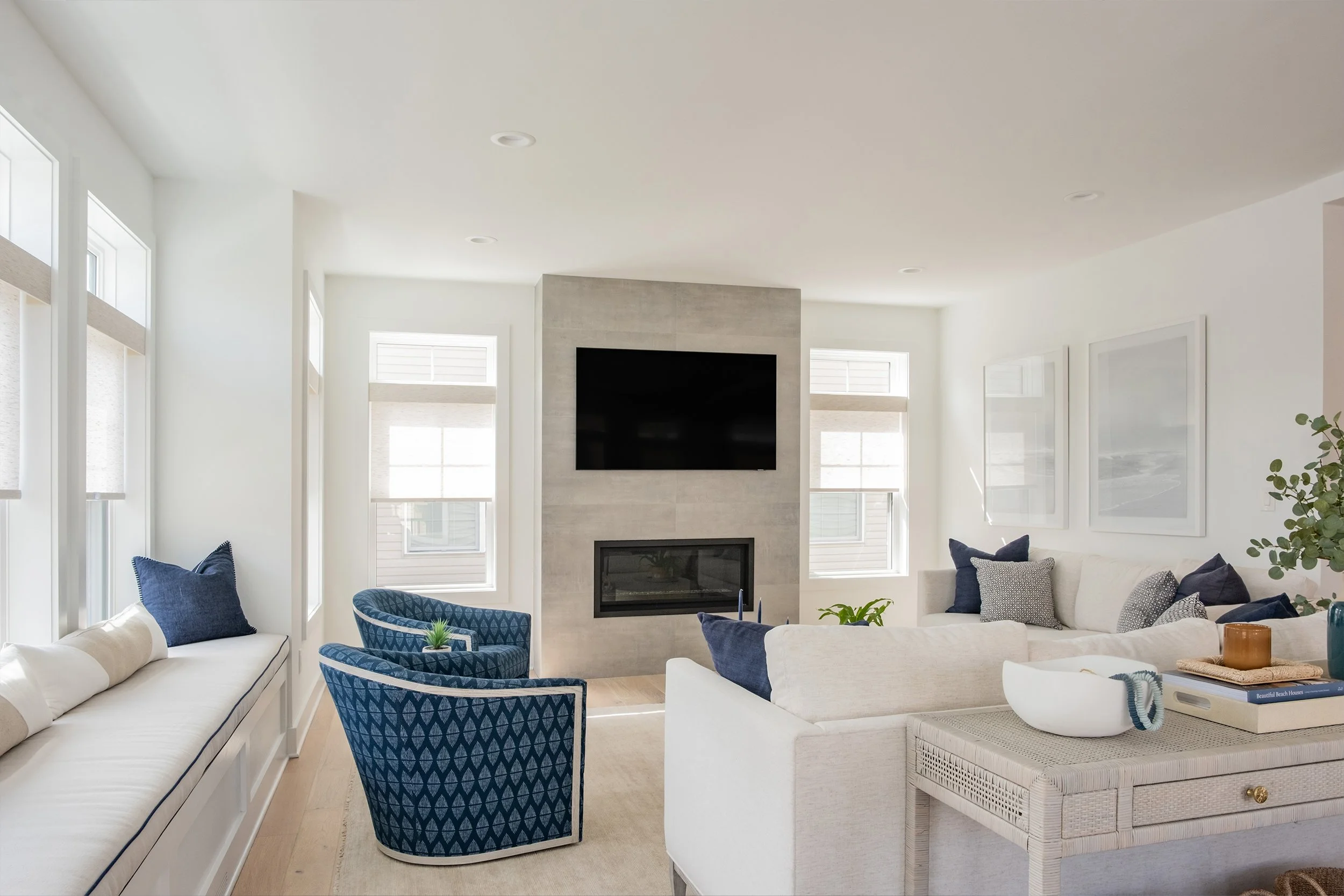 Bright living room with white walls, a fireplace, and a wall-mounted TV. Features a white sectional sofa with dark blue pillows, two blue patterned armchairs, and large windows with beige shades.