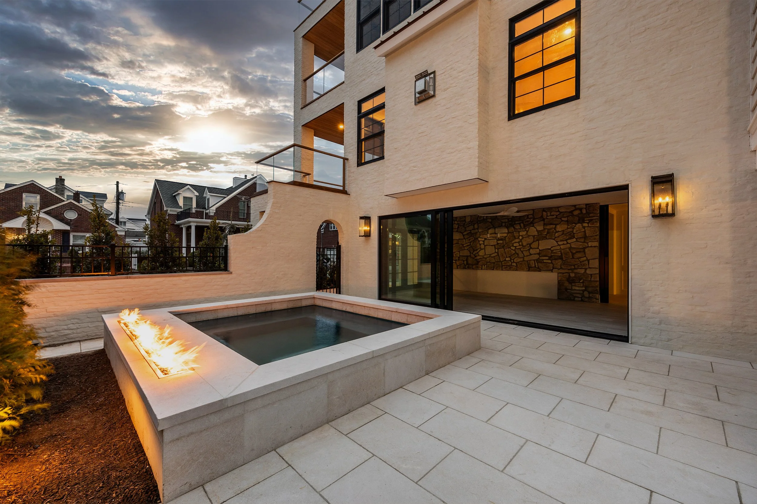Modern residential outdoor patio with a built-in water feature and fire element, large sliding glass door leading inside, stone and brick exterior, and a view of neighboring houses at sunset.