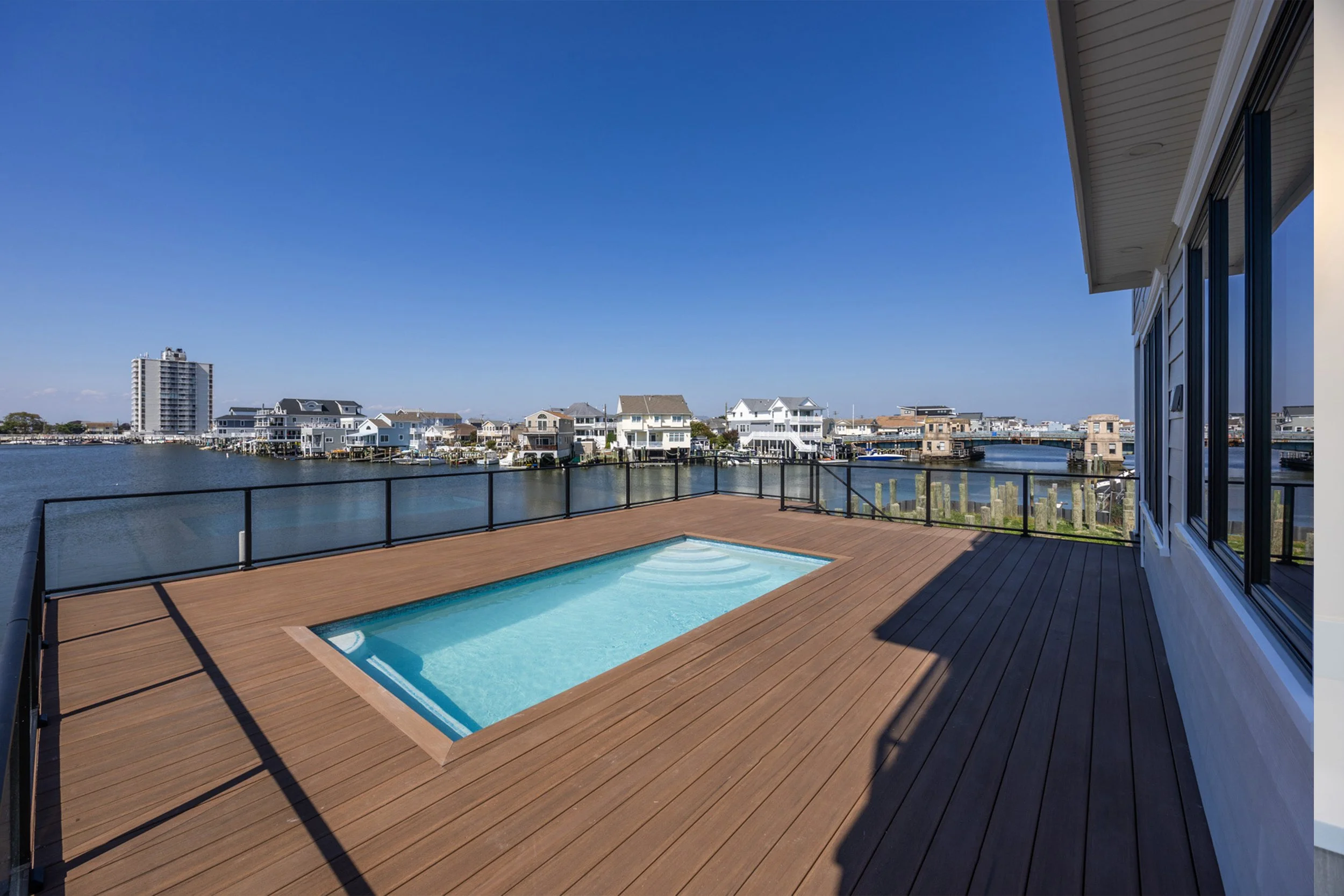 Empty rooftop hot tub on a wooden deck with a water view of houses and a canal under a clear blue sky.