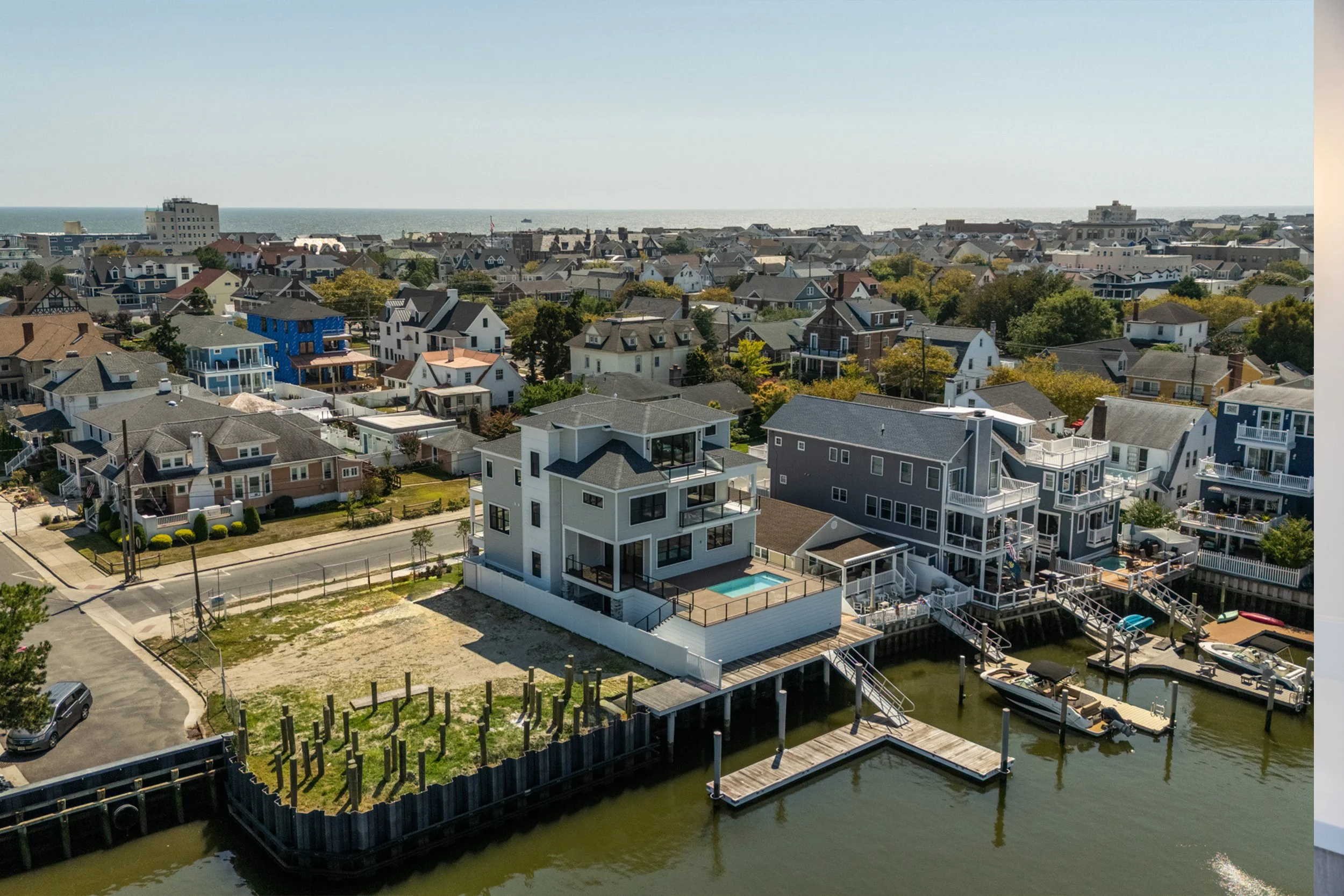 Aerial view of a coastal neighborhood with houses, boats, and a large white house with a pool, near the water.