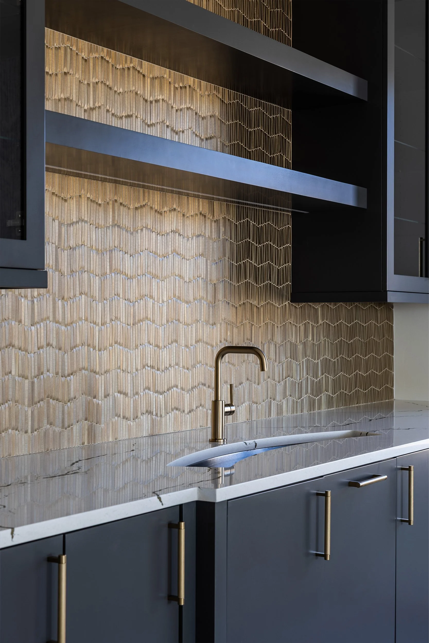 Modern kitchen with a white marble countertop, brass faucet, dark blue cabinets with brass handles, black upper cabinets, and a decorative beige tile backsplash.