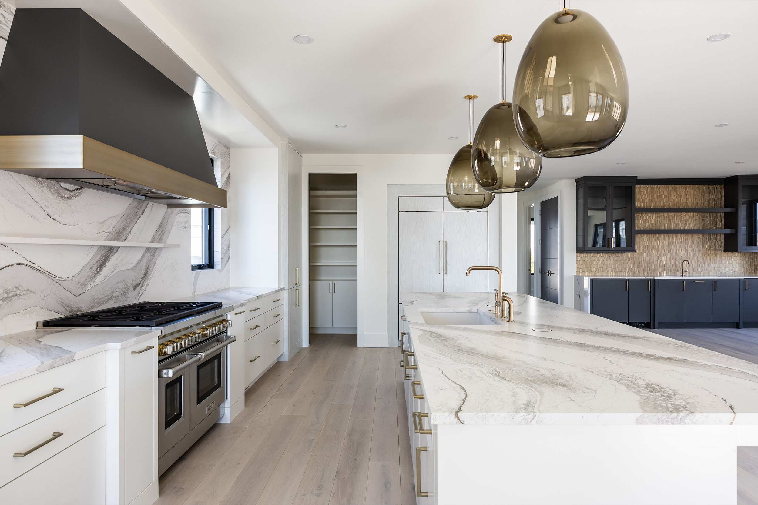 Modern kitchen with white cabinets, marble countertops, black and gold accents, and three large beige pendant lights hanging above the island.