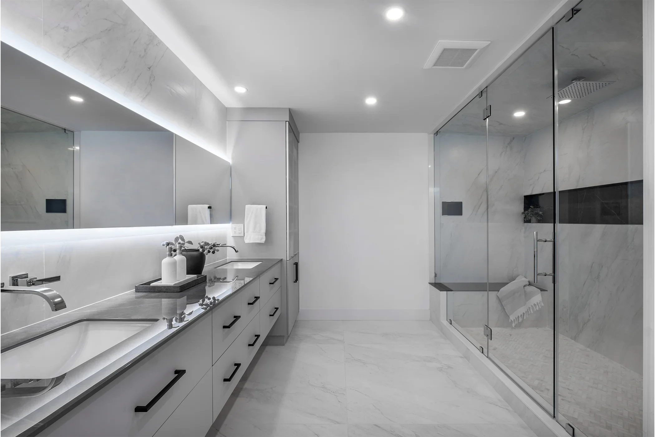 Modern bathroom with double vanity sinks, large mirror with lighting, towel rails, and a walk-in shower with glass doors, marble walls, and a built-in bench.