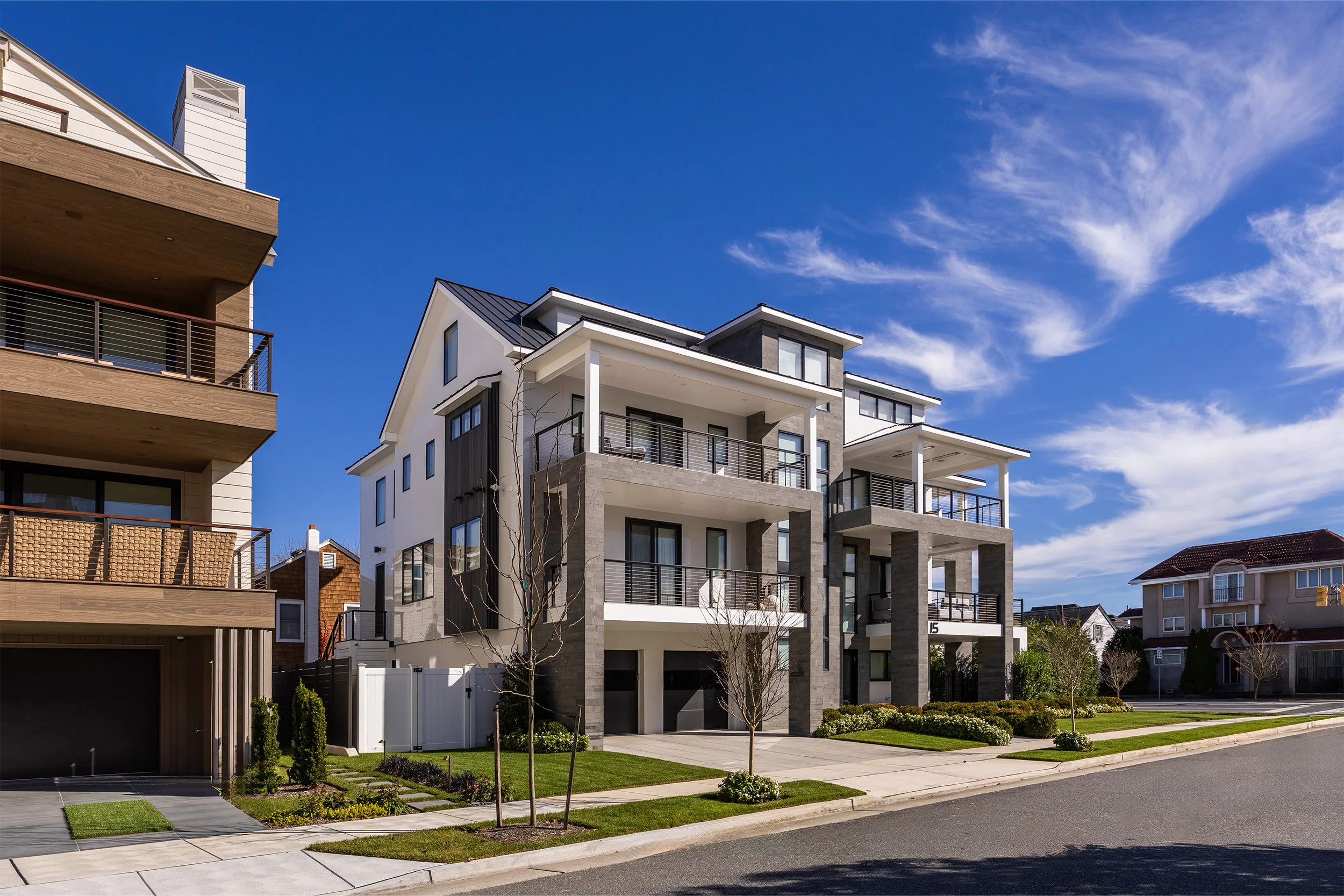 Modern multi-story residential building with balconies, white and gray facade, and landscaped front yard on a sunny day with blue sky.