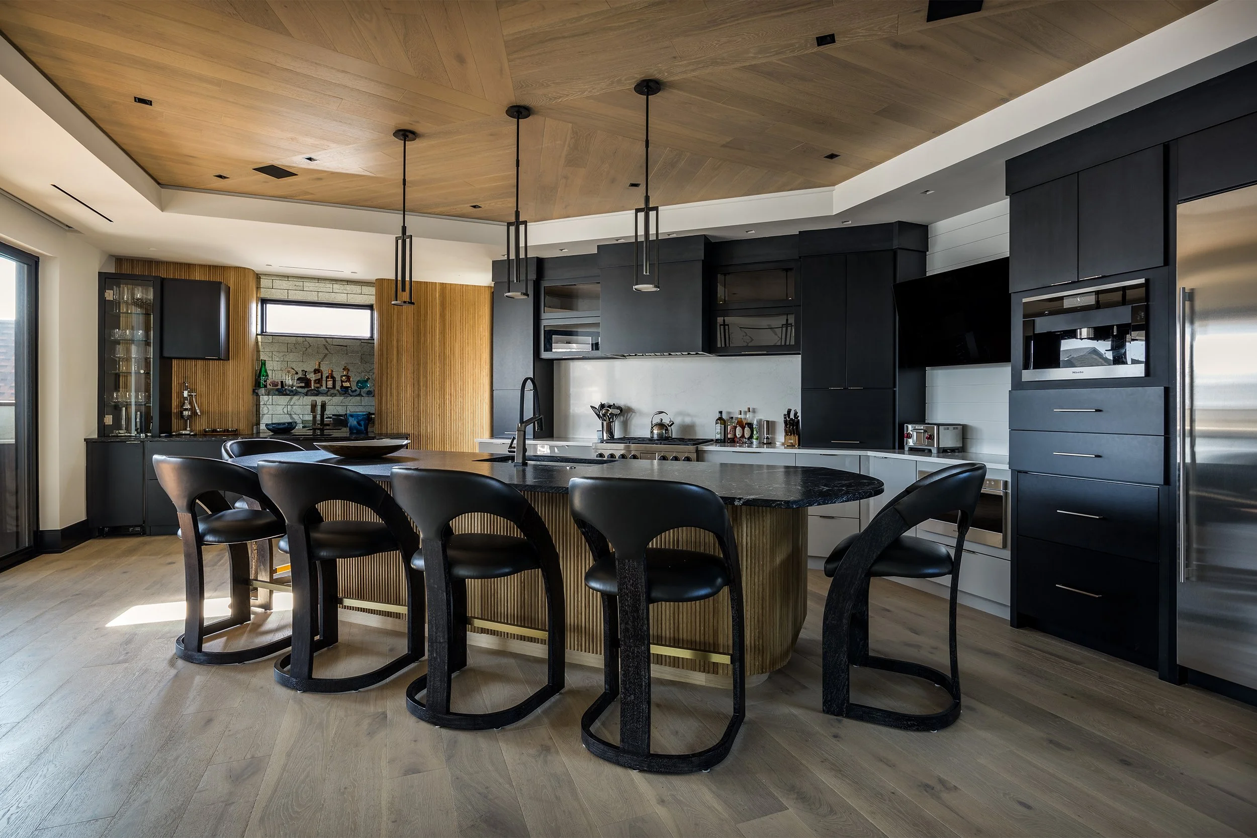Modern kitchen with black, white, and wood finishes, featuring a large island with a dark countertop, black chairs, black cabinetry, and stainless steel appliances.