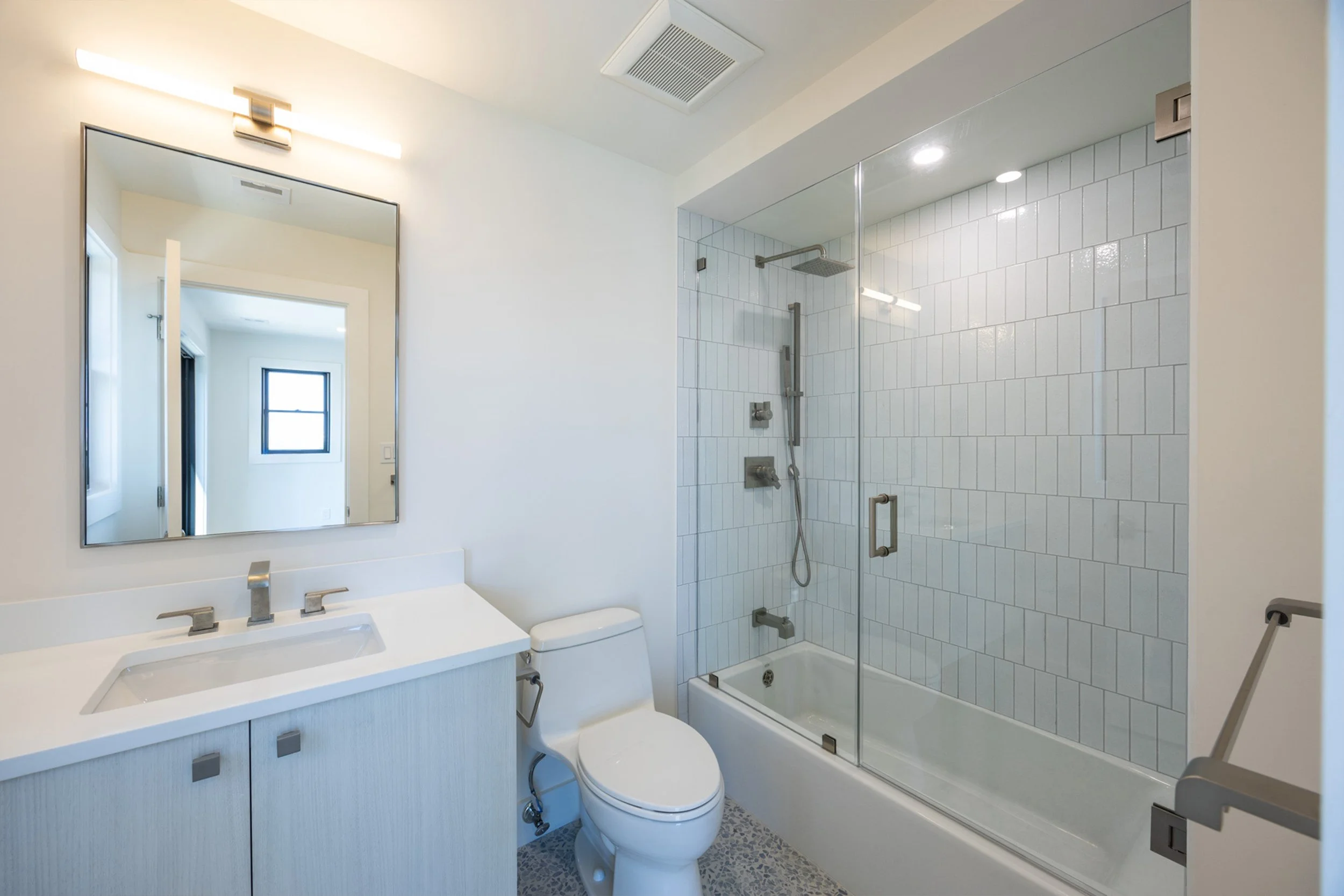 A modern bathroom featuring a large vanity with a rectangular sink, a wall-mounted mirror, a toilet, and a glass-enclosed shower with white tiles and modern fixtures.