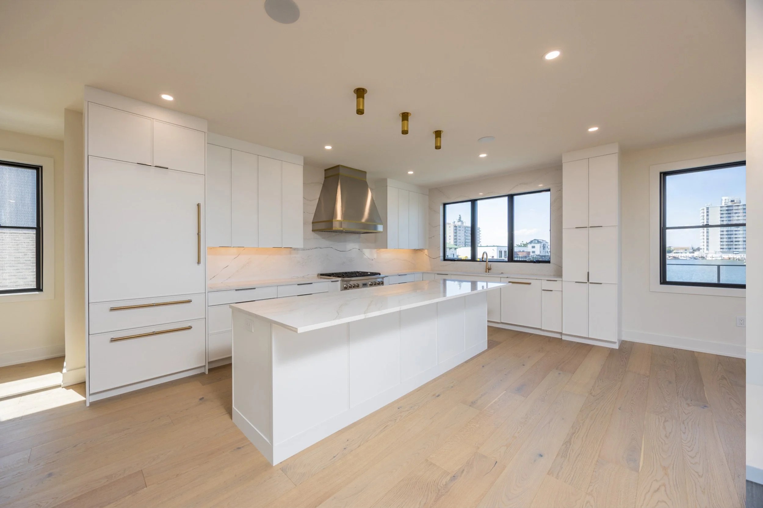 Modern white kitchen with marble countertops, sink, and large windows overlooking a cityscape.