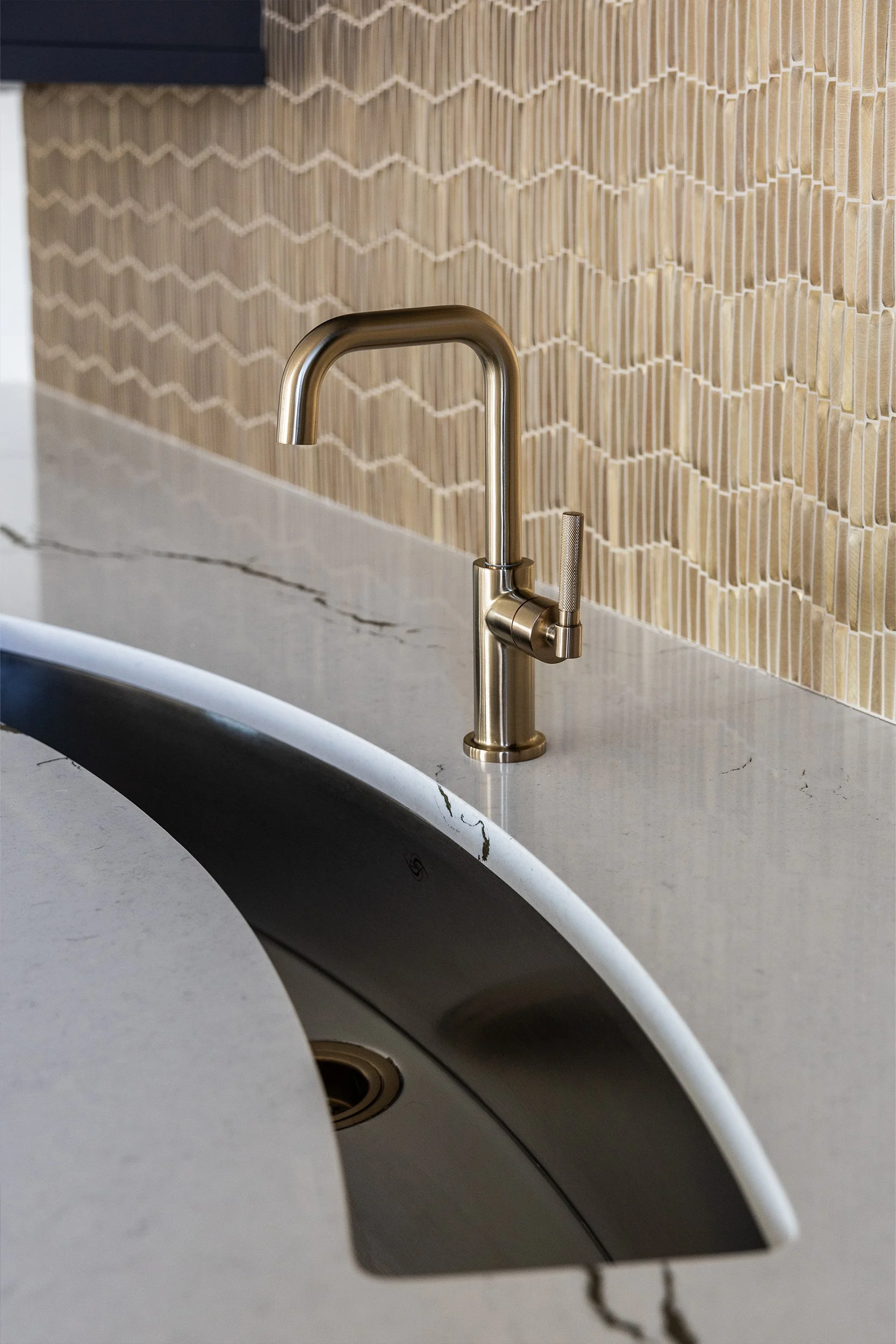 Close-up of a modern metallic kitchen faucet above a white marble countertop with a curved edge, in front of a beige tile backsplash with a textured, geometric pattern.