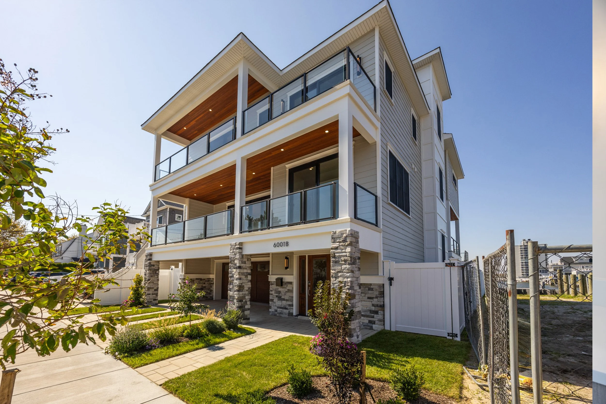 A modern three-story residential building with white siding, stone accents, and glass balconies. Well-maintained front yard with grass and small shrubs, sidewalk, and a white fence on the side.