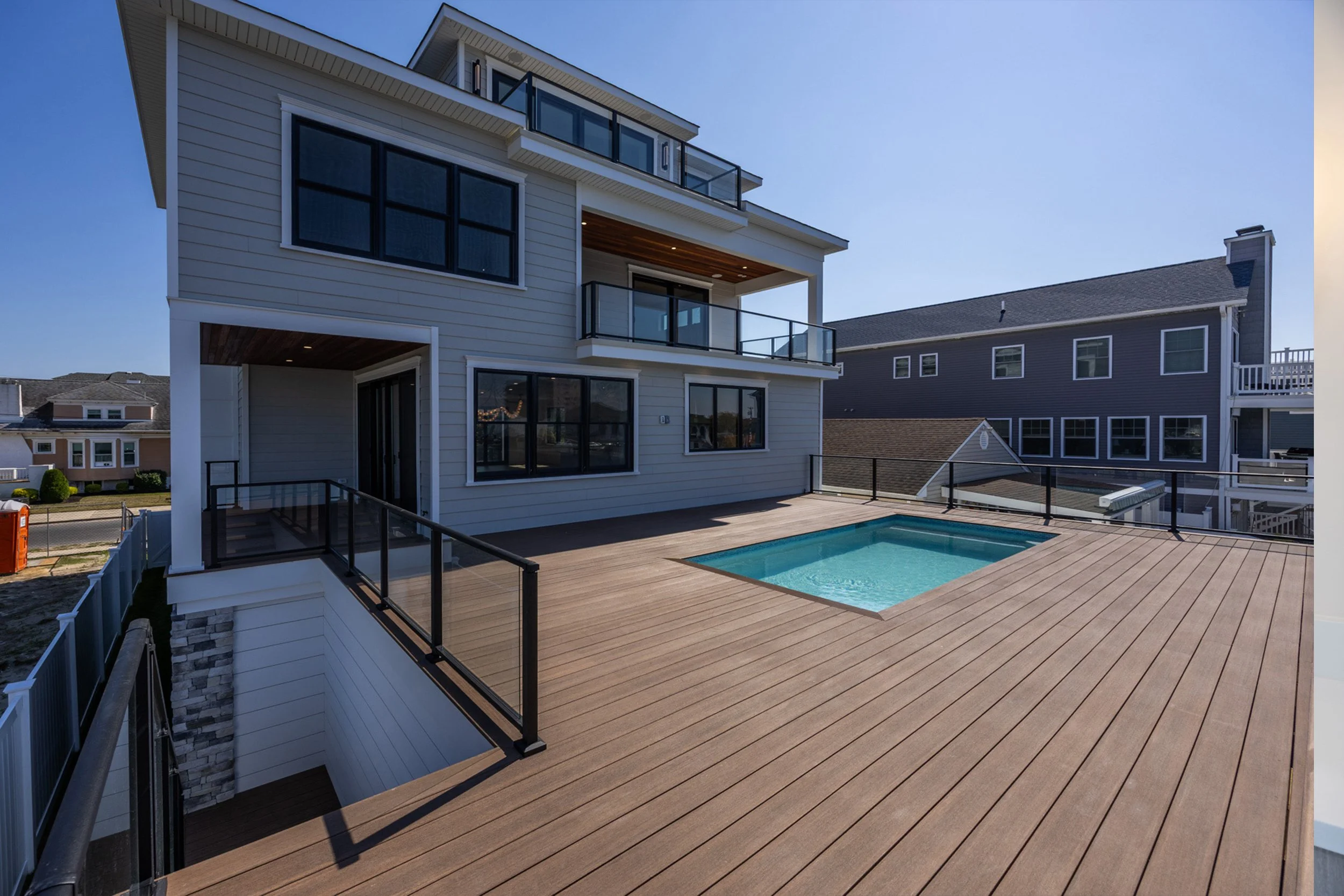 A modern house with a spacious wooden deck, a small rectangular swimming pool, and multiple balconies with black metal railings under a clear blue sky.