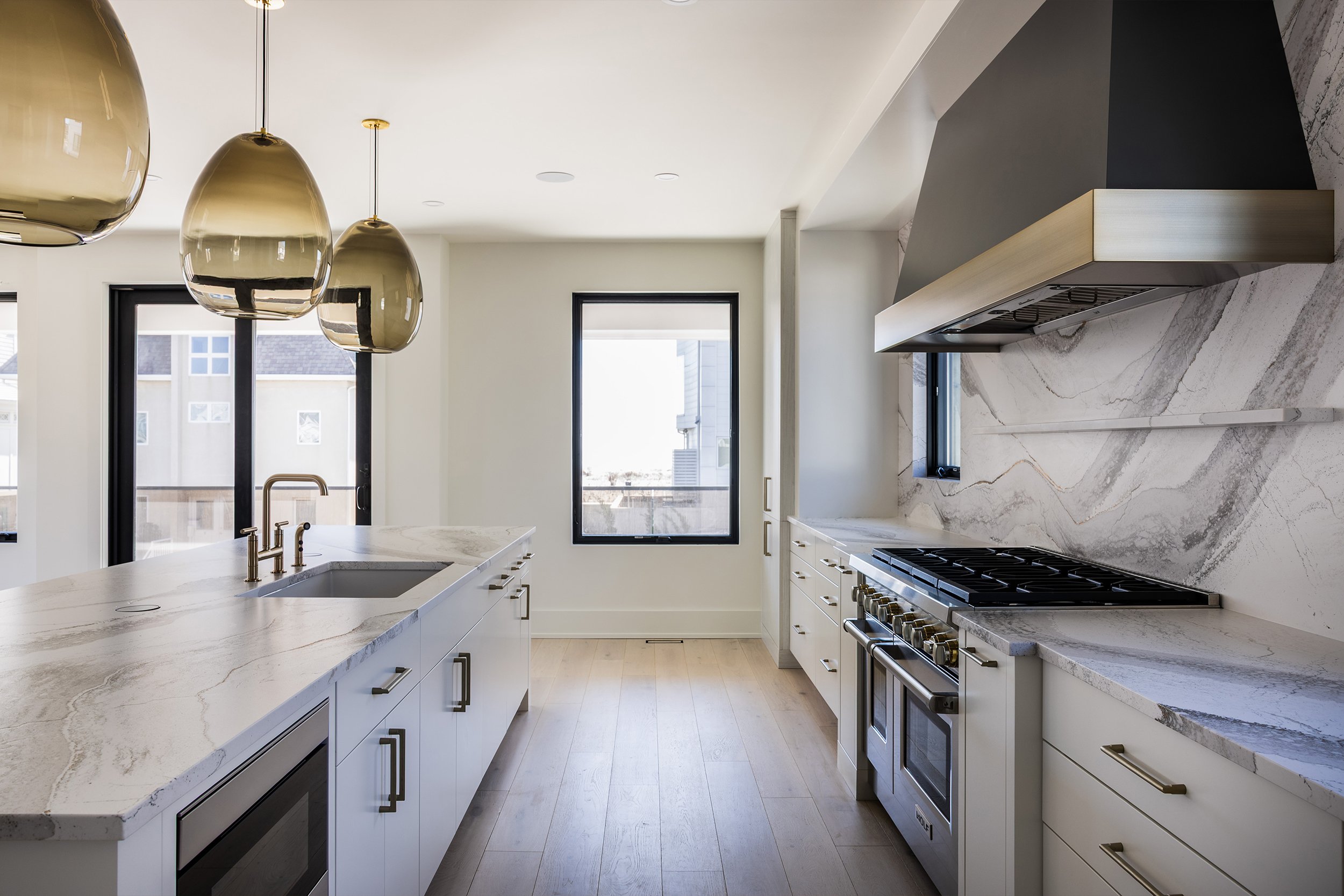 Modern kitchen with white cabinetry, marble countertops, stainless steel appliances, black-framed windows, and three gold pendant lights.