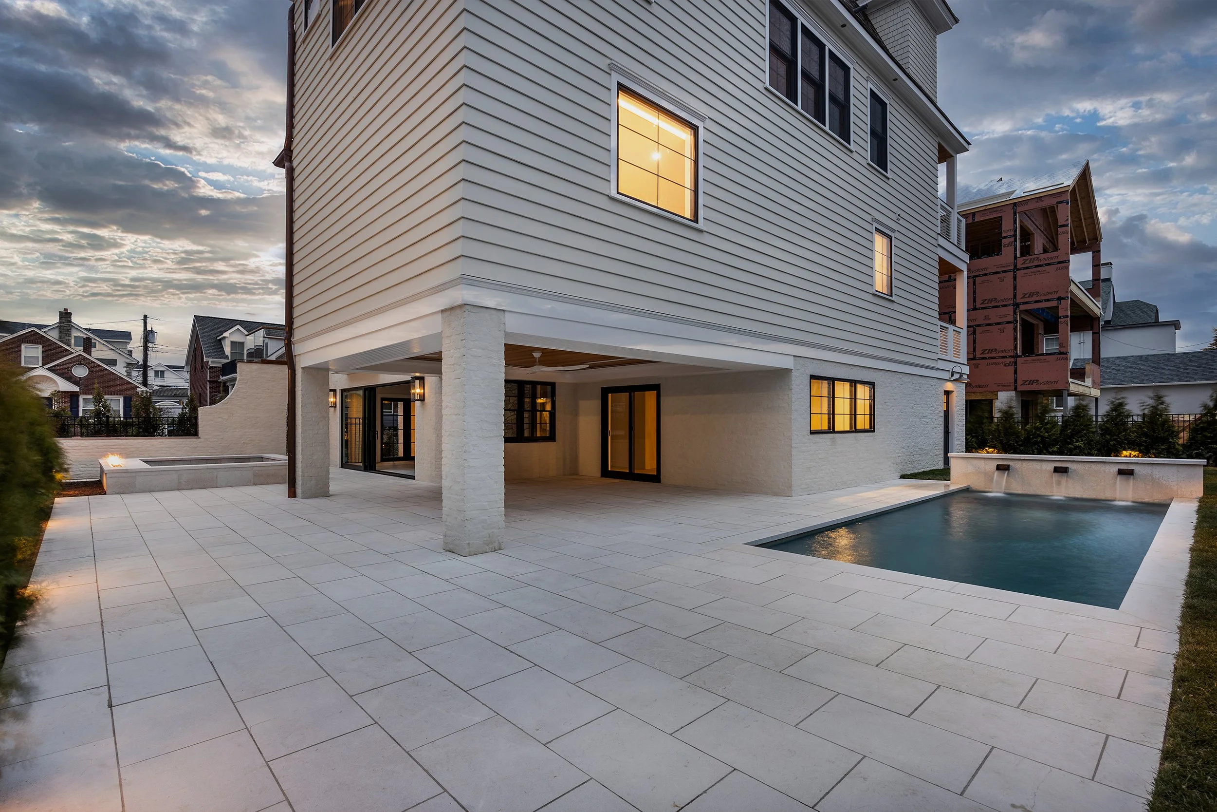 Modern house with white siding and stone accents, backyard patio with pool, and large windows with warm interior lighting, under a partly cloudy sky at sunset.