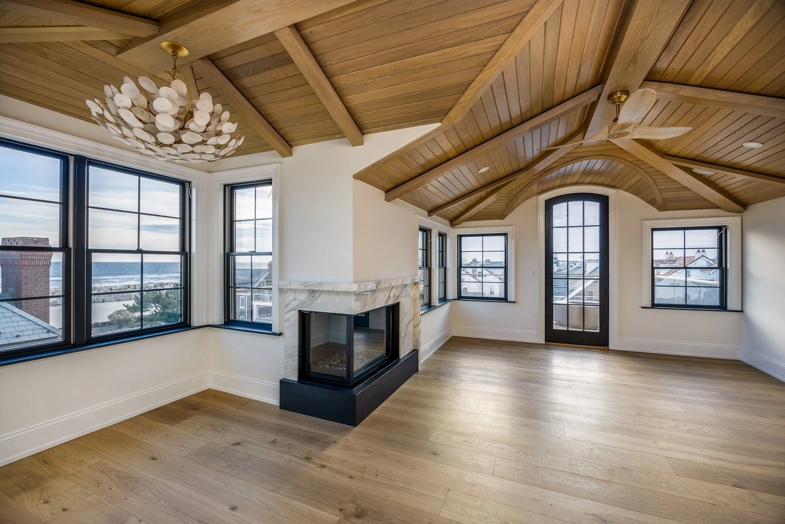 Bright room with large windows showing an ocean view, a wooden vaulted ceiling with a ceiling fan, a white marble fireplace, and light hardwood flooring.
