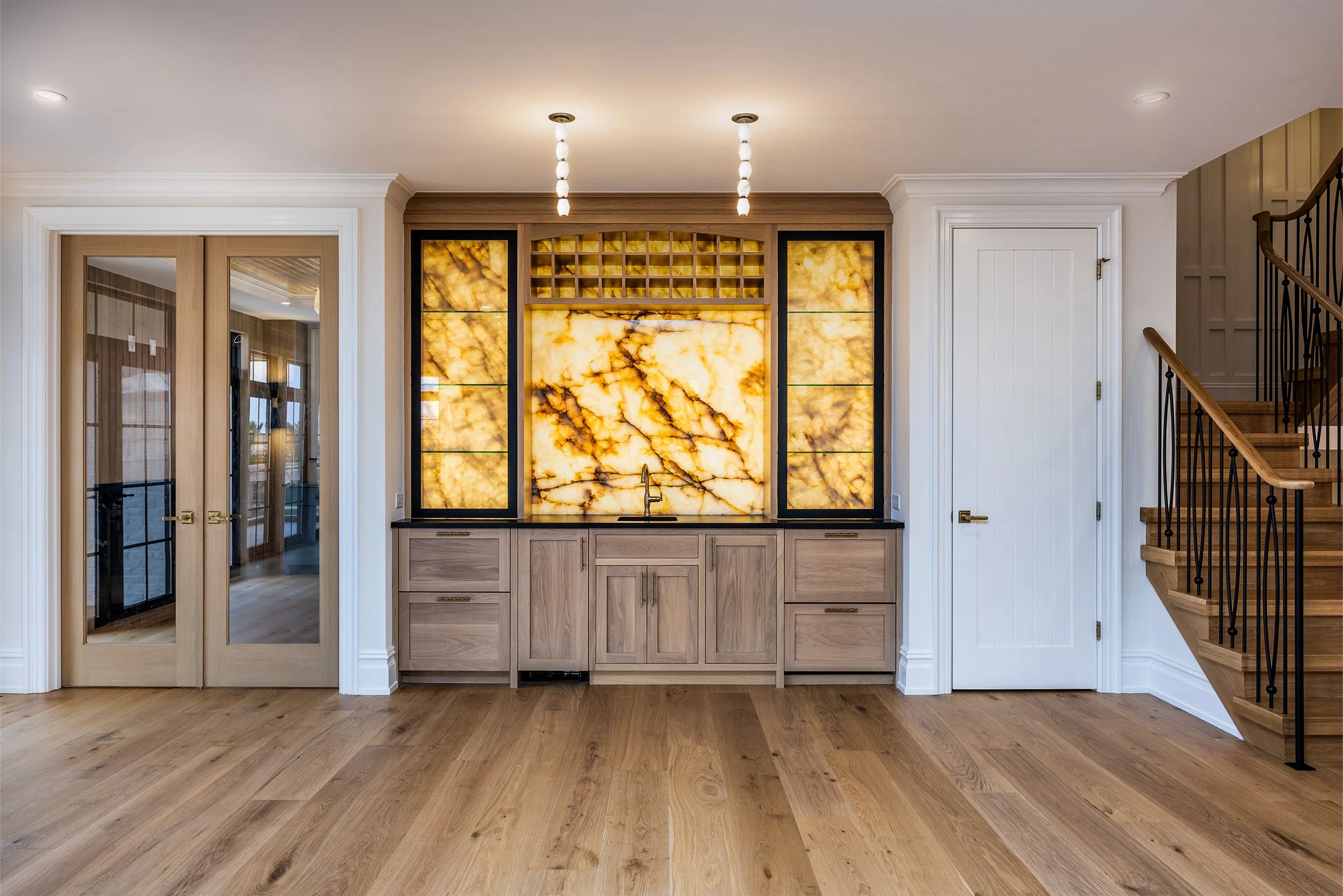 Interior of a home with a built-in bar featuring a backlit onyx wall, light wood cabinetry, and a black countertop. To the left, there are glass-paneled double doors, and to the right, a staircase with wooden steps and black metal railing. The floor is made of wide light wood planks, and the ceiling has recessed and decorative lighting.