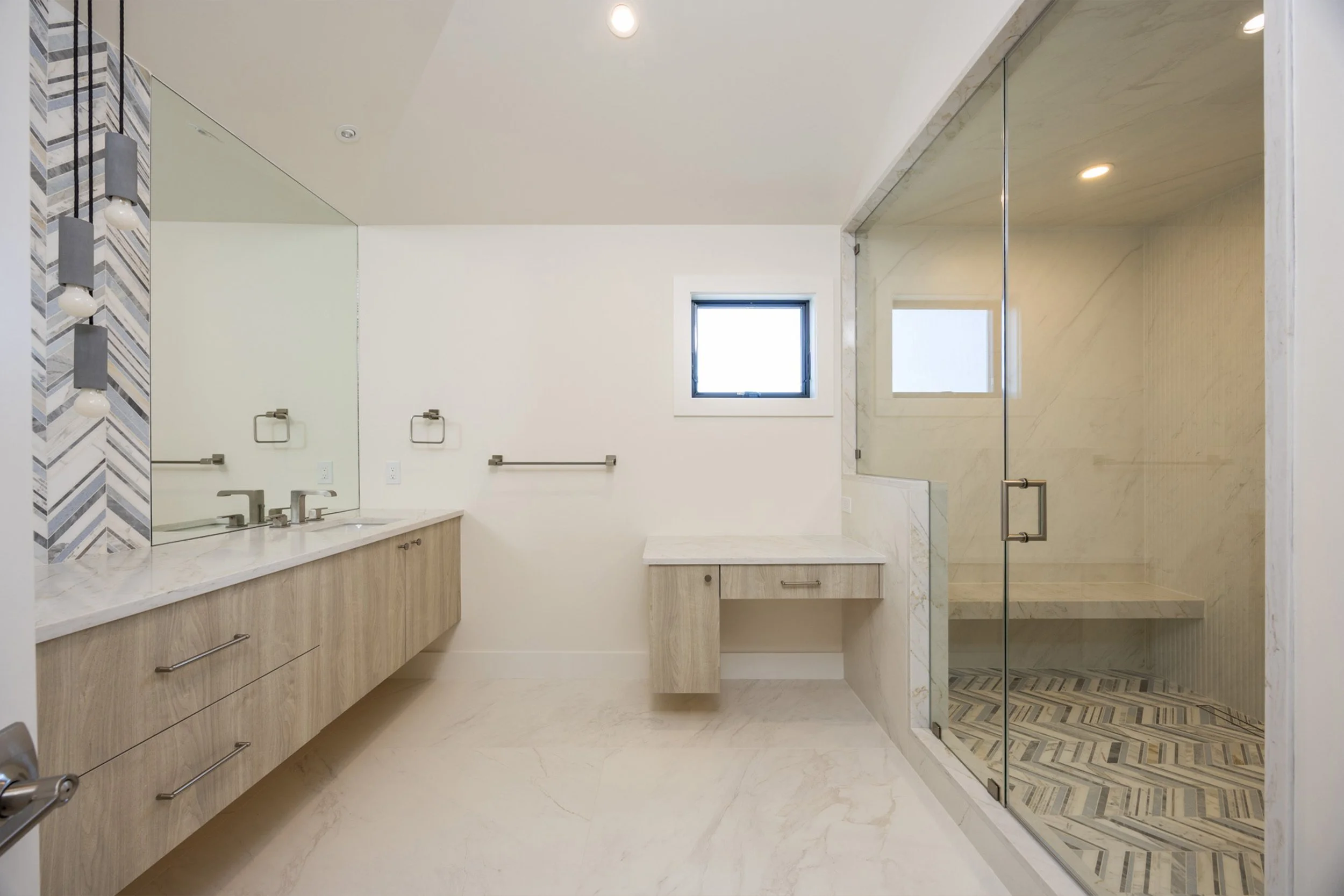 Modern bathroom with marble countertops, wooden cabinetry, large mirror, small square window, and glass-enclosed shower with patterned tile floor.