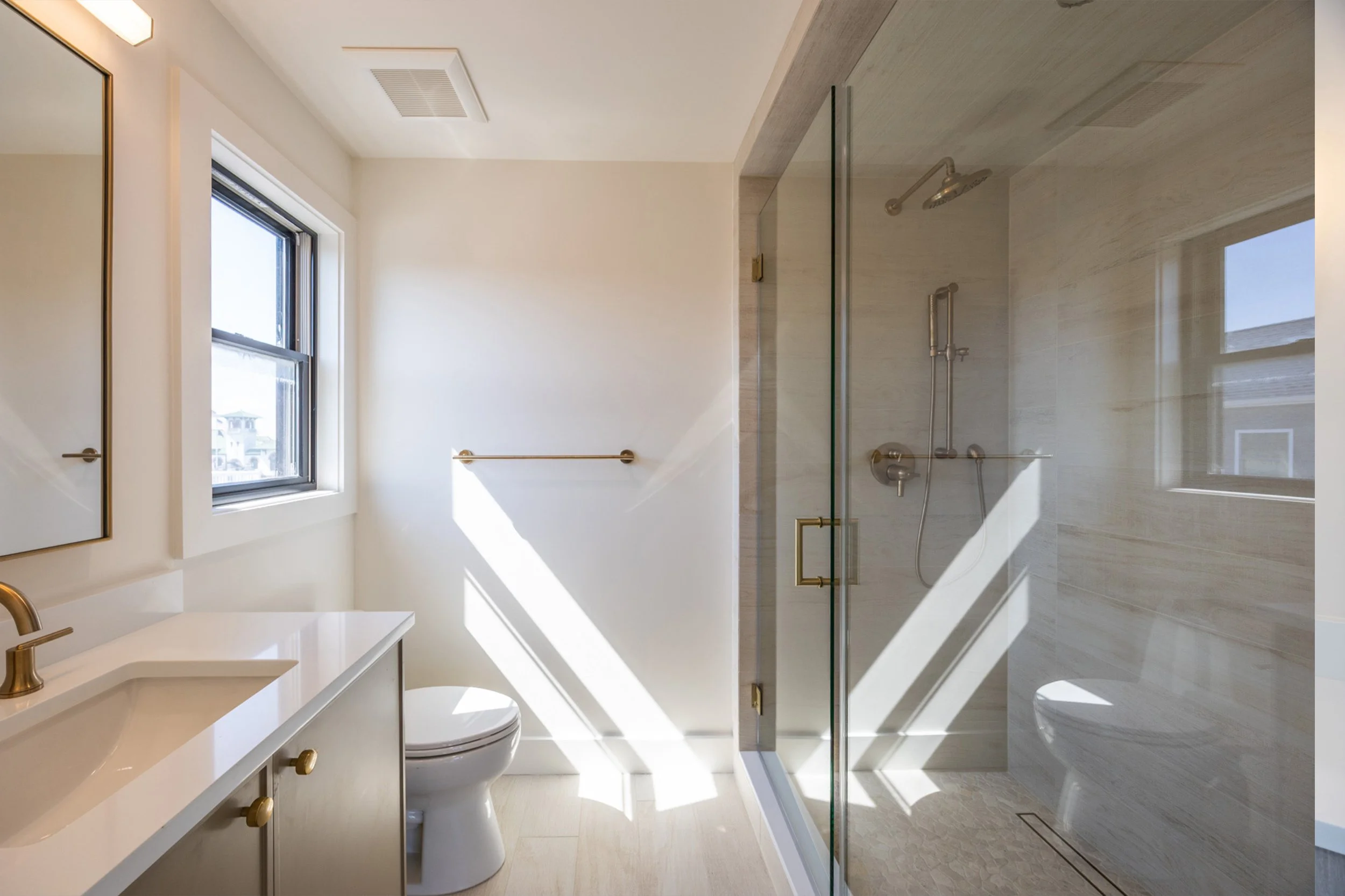 Modern bathroom with white walls, a window, a mirror, a vanity with gold handles, a toilet, and a walk-in shower with a glass door and beige marble walls.