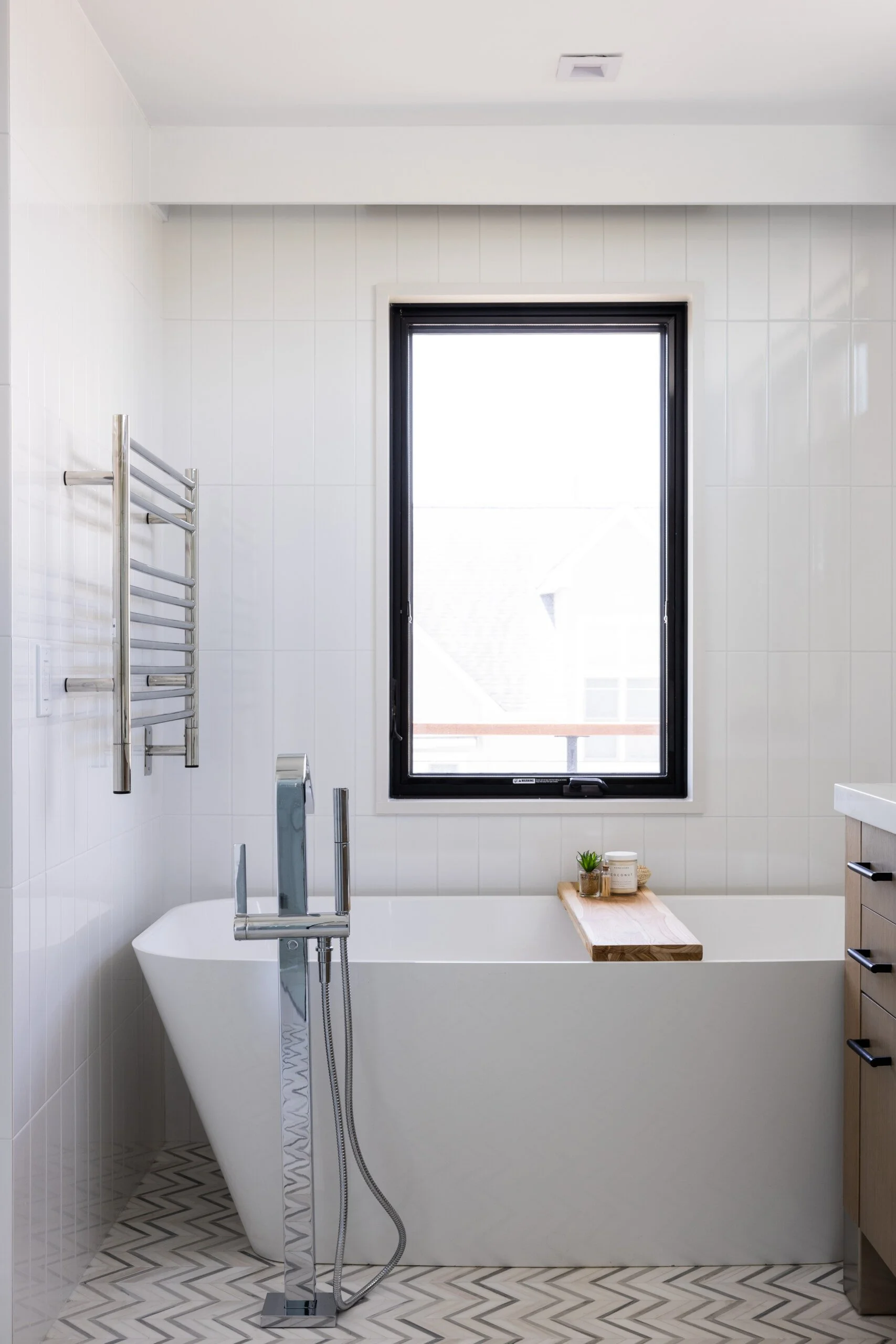 Modern bathroom with a freestanding bathtub, large window, wall-mounted towel warmer, and wooden shelf holding toiletries and decorative items.