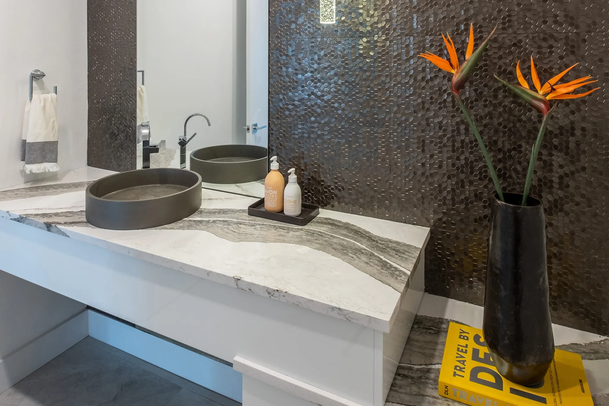 Modern bathroom vanity with a white marble countertop, dark round vessel sinks, a large mirror, bottles of lotion, a tall black vase with orange and green flowers, and a yellow magazine on the floor.
