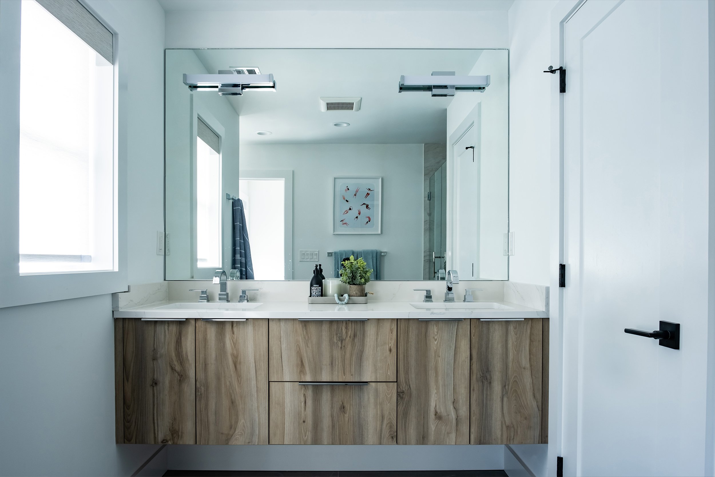 Modern bathroom with double vanity and large mirror, white countertops, wooden cabinets, black fixtures, and natural light from windows.