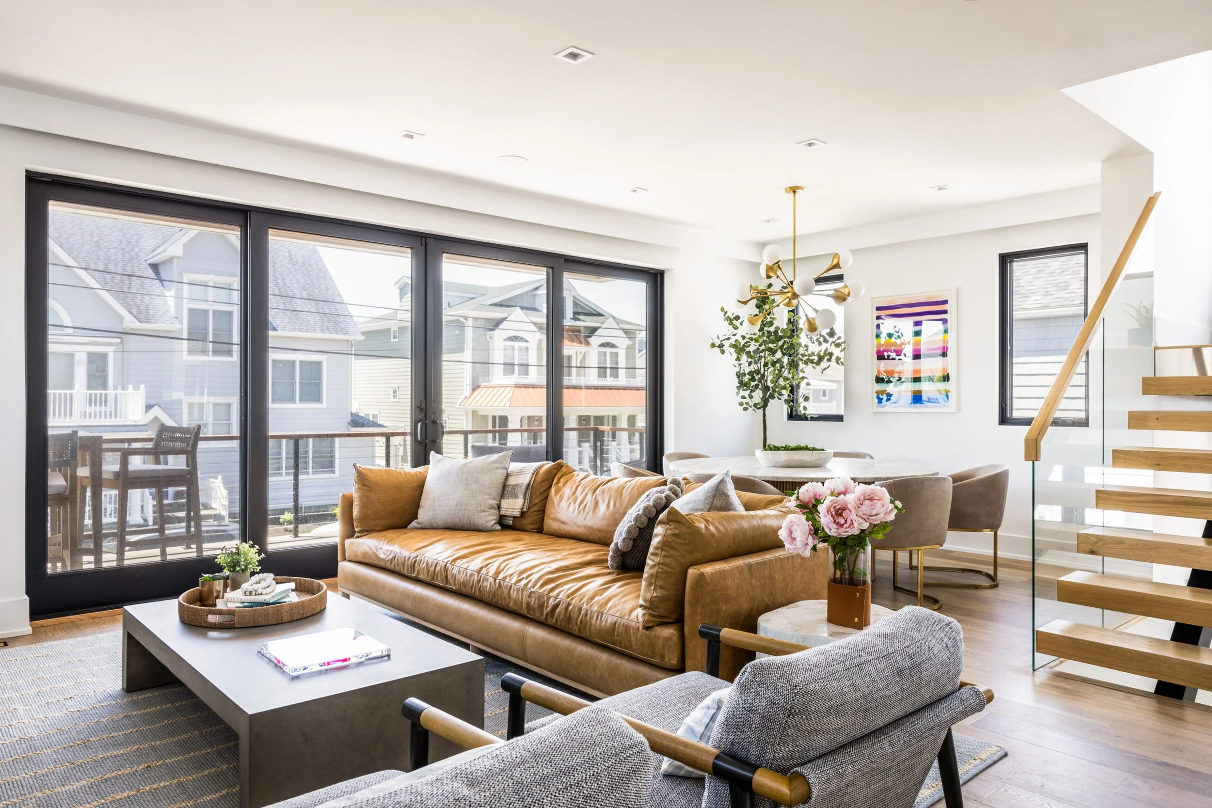 Modern living room with tan leather sofa, gray armchair, coffee table, large sliding glass door, staircase with wooden steps, artwork on the wall, pink flowers, potted plant, and a dining area in the background.