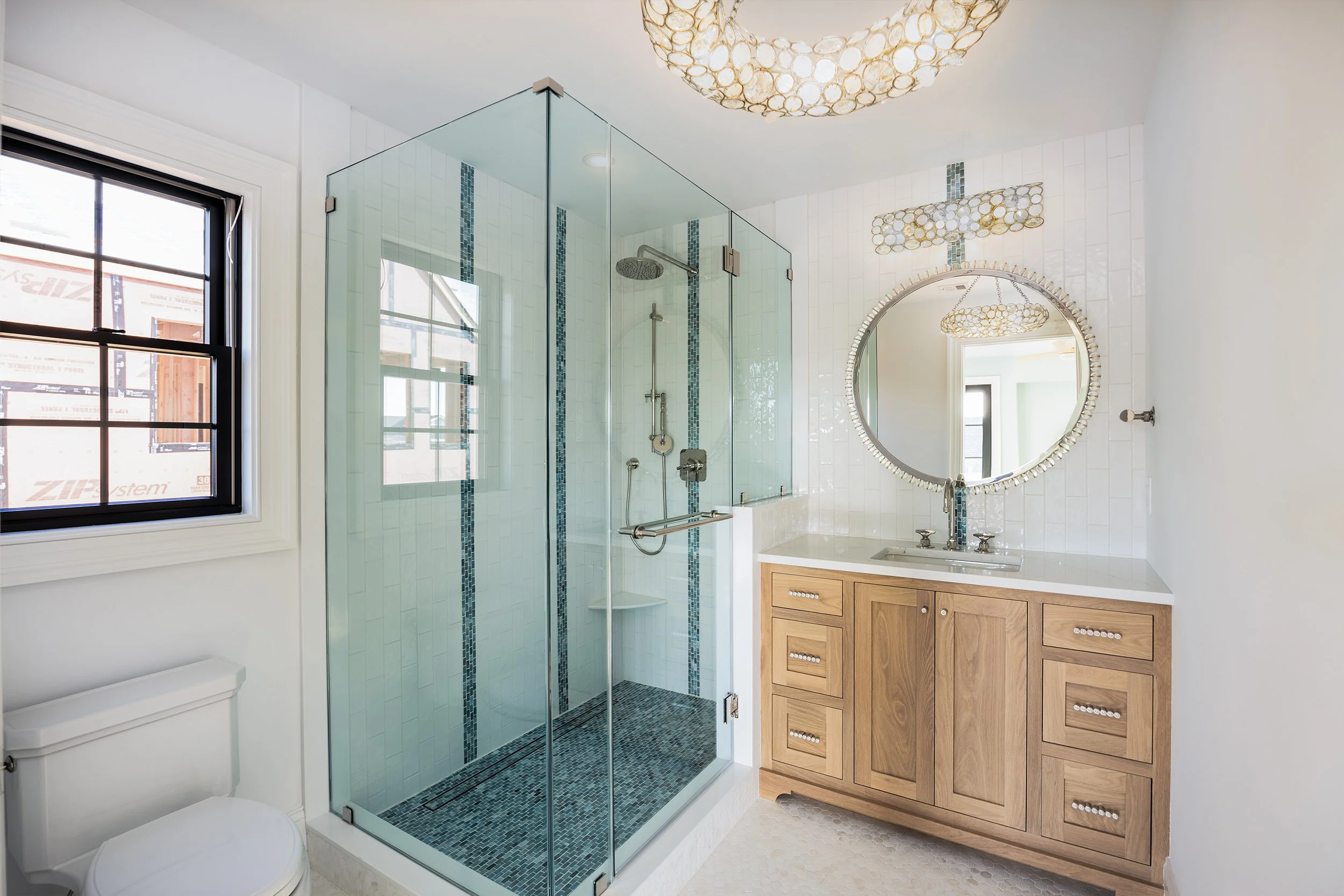 Modern bathroom with a glass-enclosed shower, wooden vanity with a round mirror, and a window allowing natural light.