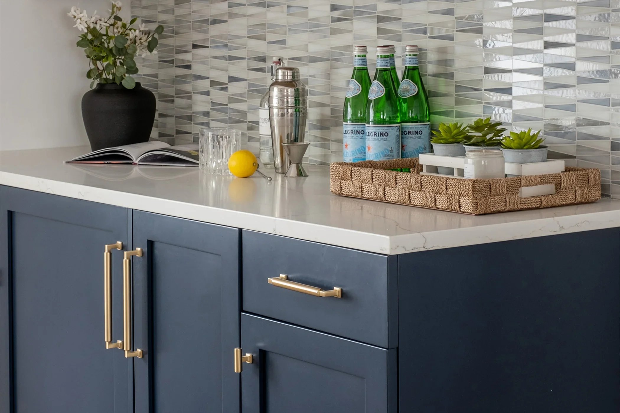Kitchen countertop with blue cabinets, gold handles, a black plant pot with greenery, an open book, a glass, lemon, bar tools, three bottles of sparkling water, and a tray with succulents and small containers.