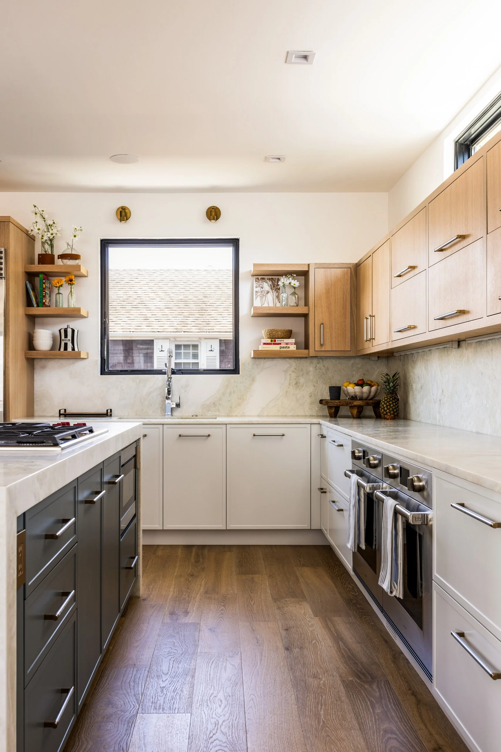 Modern kitchen with wooden and white cabinets, marble countertops, a window above the sink, and open shelves with decorative items.