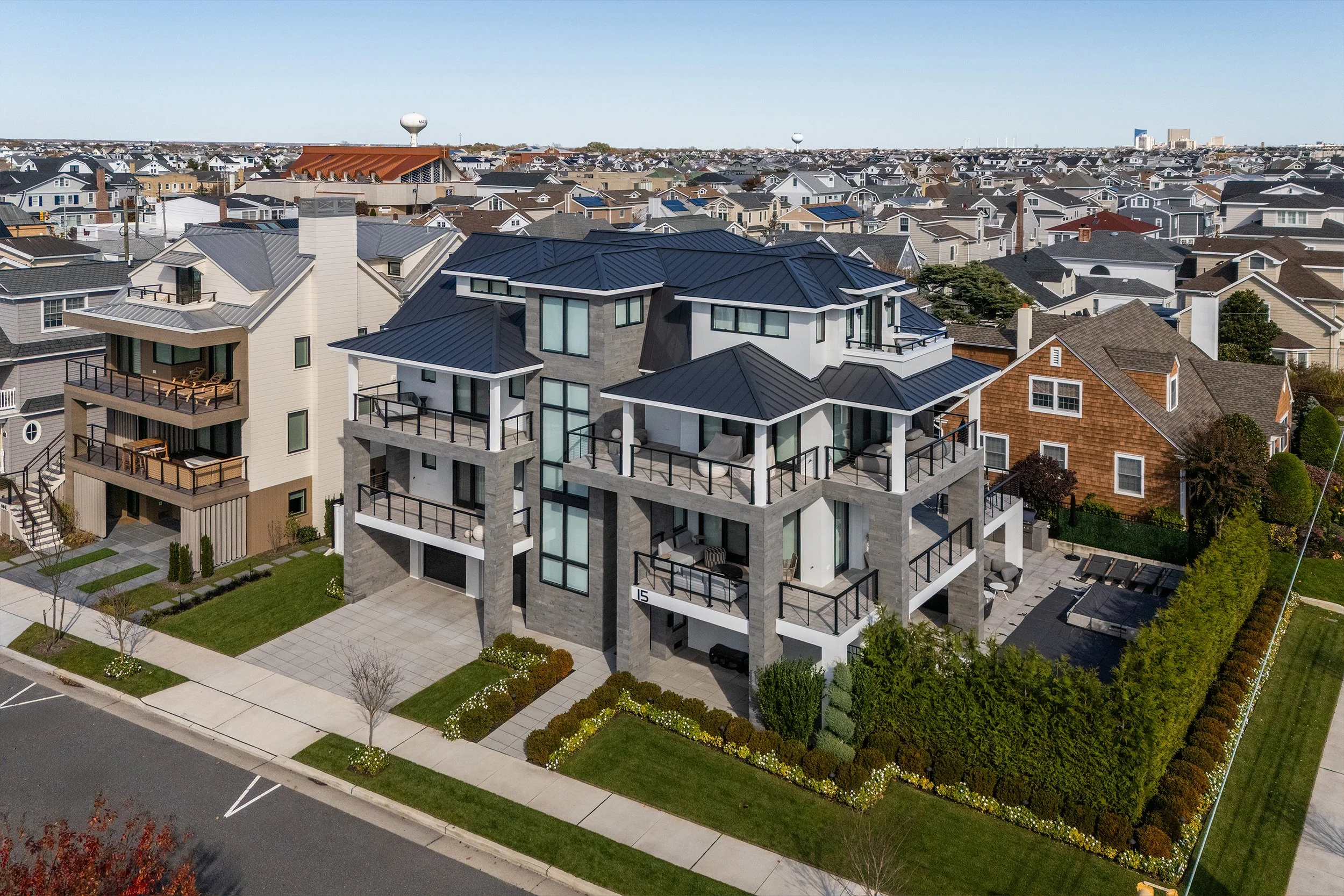 A modern multi-story residential building with gray stone walls and black metal roofs, surrounded by green lawns, trees, and a sidewalk, in a neighborhood with similar houses.