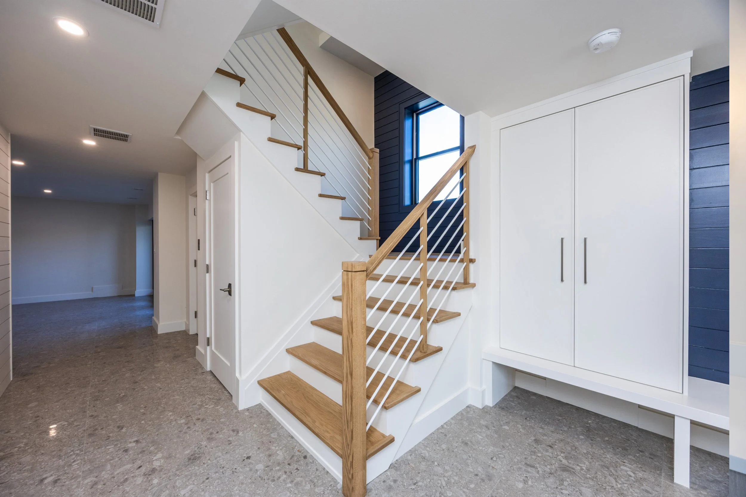 Interior view of a modern staircase with wooden steps and white railings leading to an upper floor, next to a window and a built-in white cabinet with double doors, in a well-lit living space.
