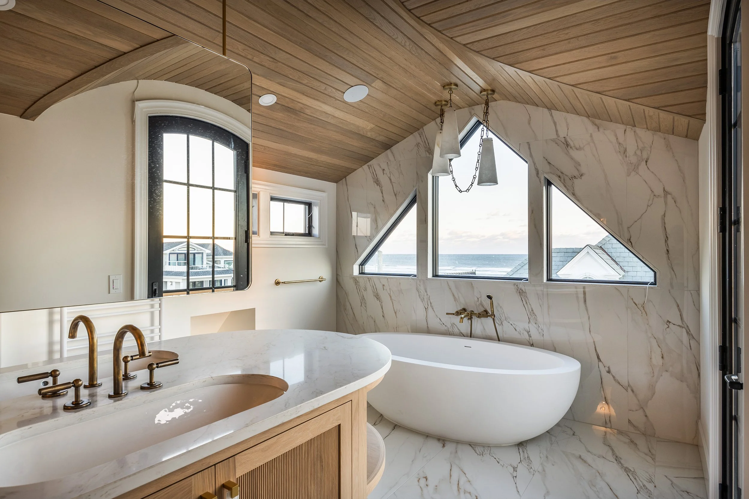Elegant bathroom with a double sink vanity, a freestanding bathtub, and large windows offering ocean views, featuring marble walls and a wooden ceiling.