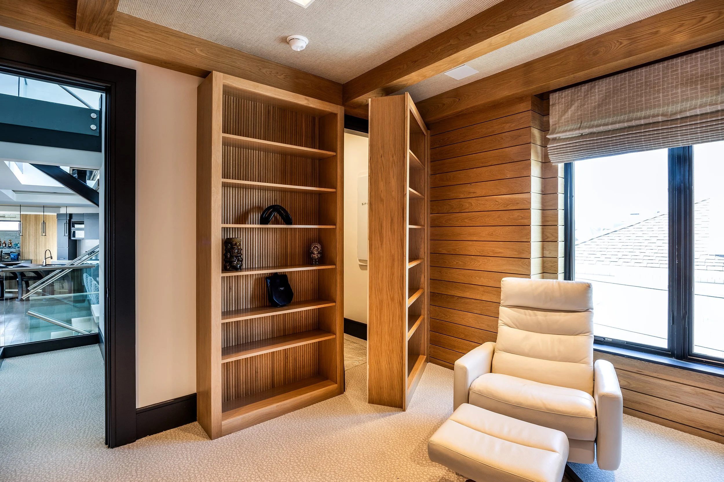 Corner of a room with wooden walls and ceiling, a white recliner chair with footrest next to a large window, and an open wooden bookshelf with a few decorative items, in a modern home interior.